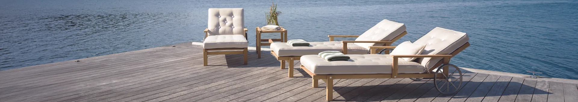Outdoor scene with a wooden dock featuring a light-colored armchair, two sun loungers with wheels, and a side table by blue water.