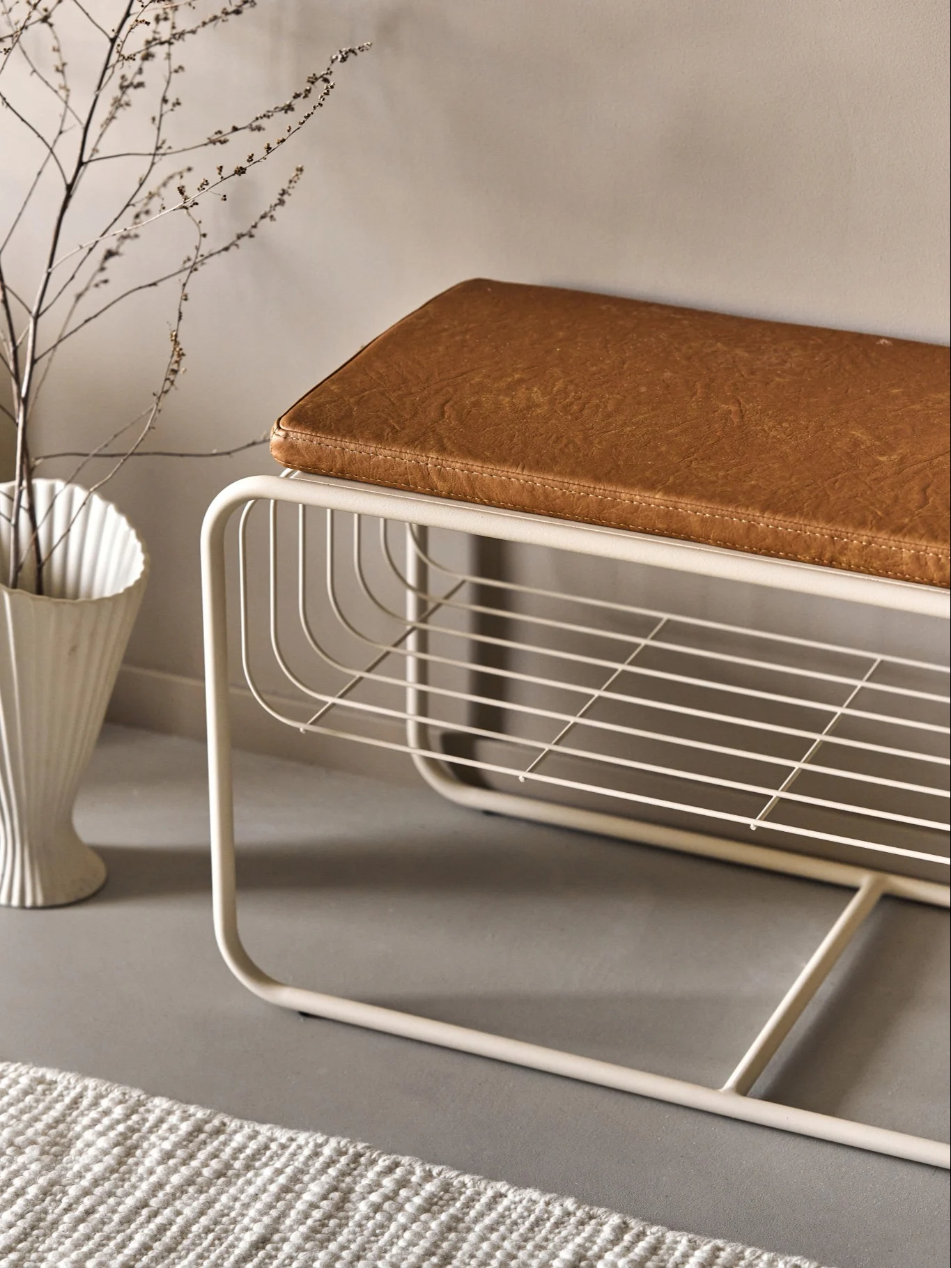 A modern bench with a light metal frame, brown cushion, and a lower shelf, next to a vase with branches and a rug.