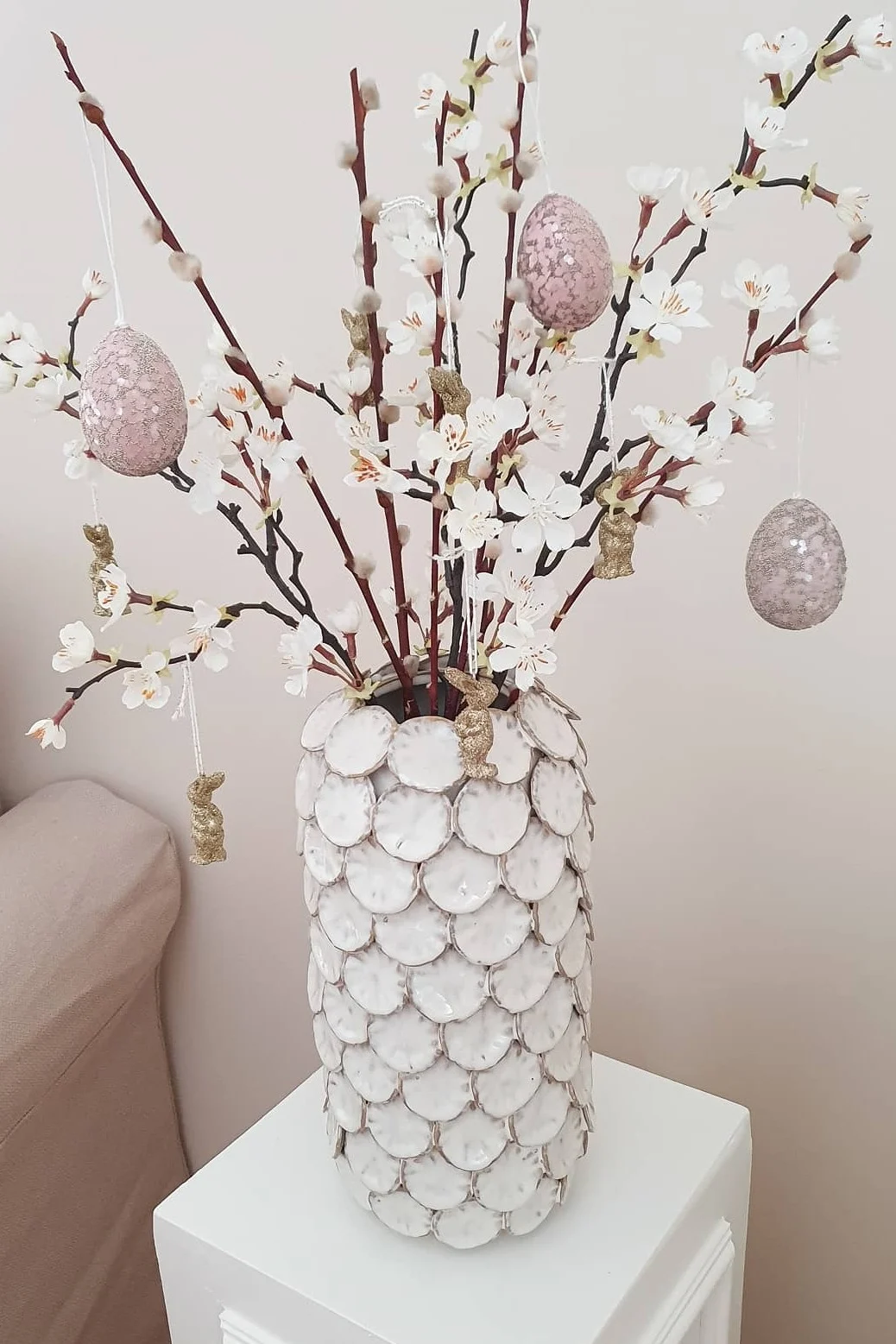 Easter decoration with a white vase, branches with white blossoms and pussy willows, pink speckled eggs, and gold glitter bunnies.