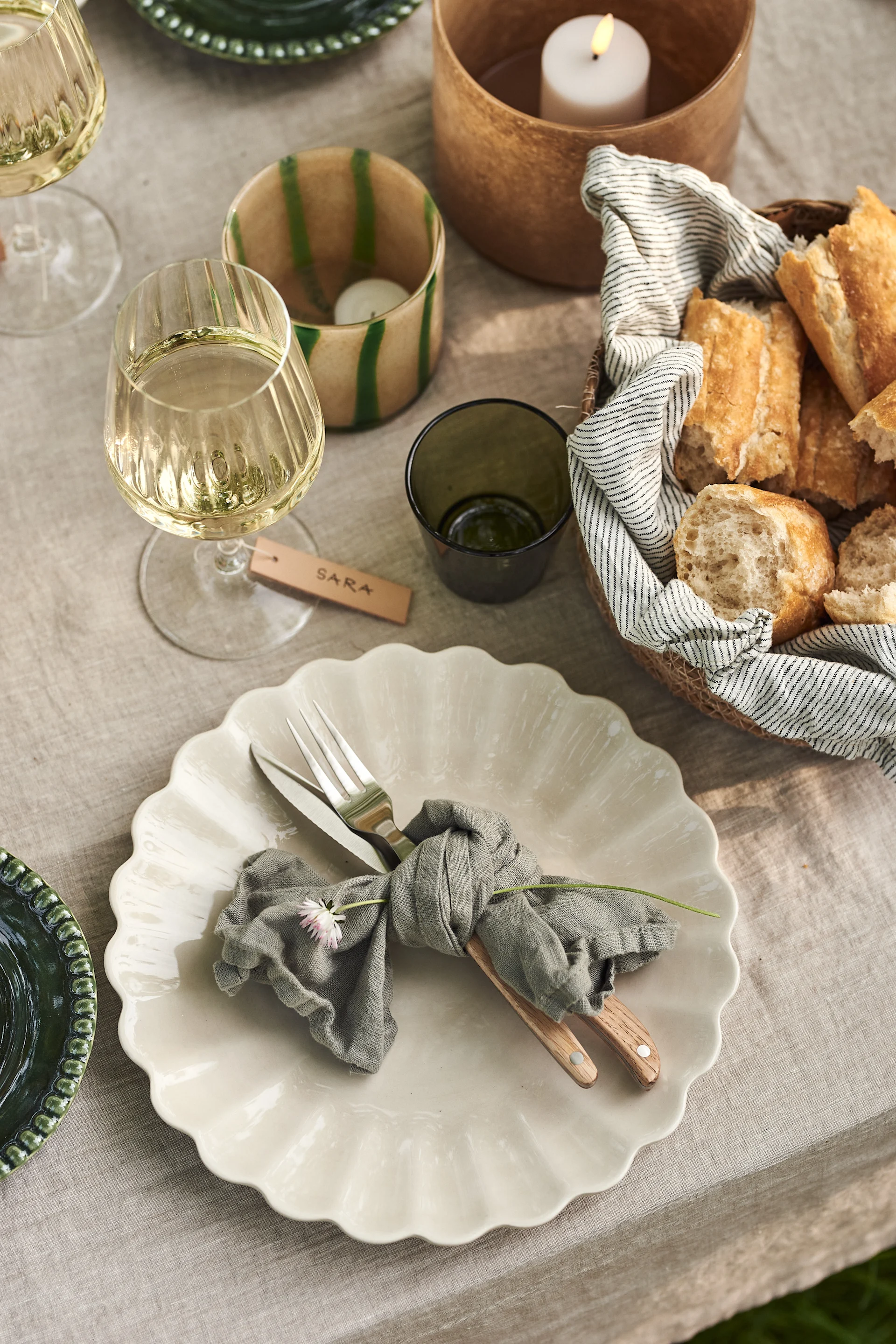 Outdoor dining table set with a white scalloped plate, cutlery tied with a gray napkin, a wine glass, and a bread basket.