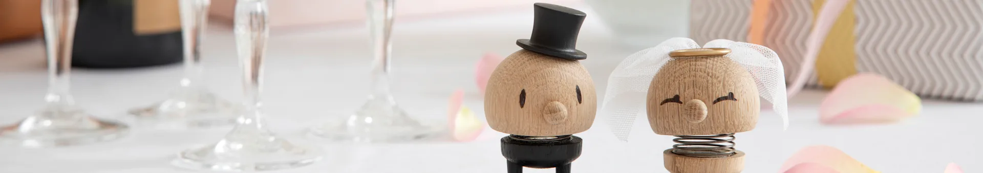 Wooden wedding figurines of a groom with a top hat and a bride with a veil, surrounded by glasses and rose petals.