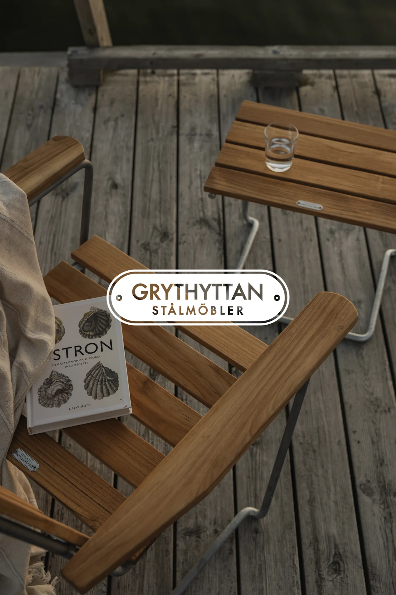 Outdoor wooden slatted chair with a blanket and a book, next to a matching table with a glass of water on a deck.