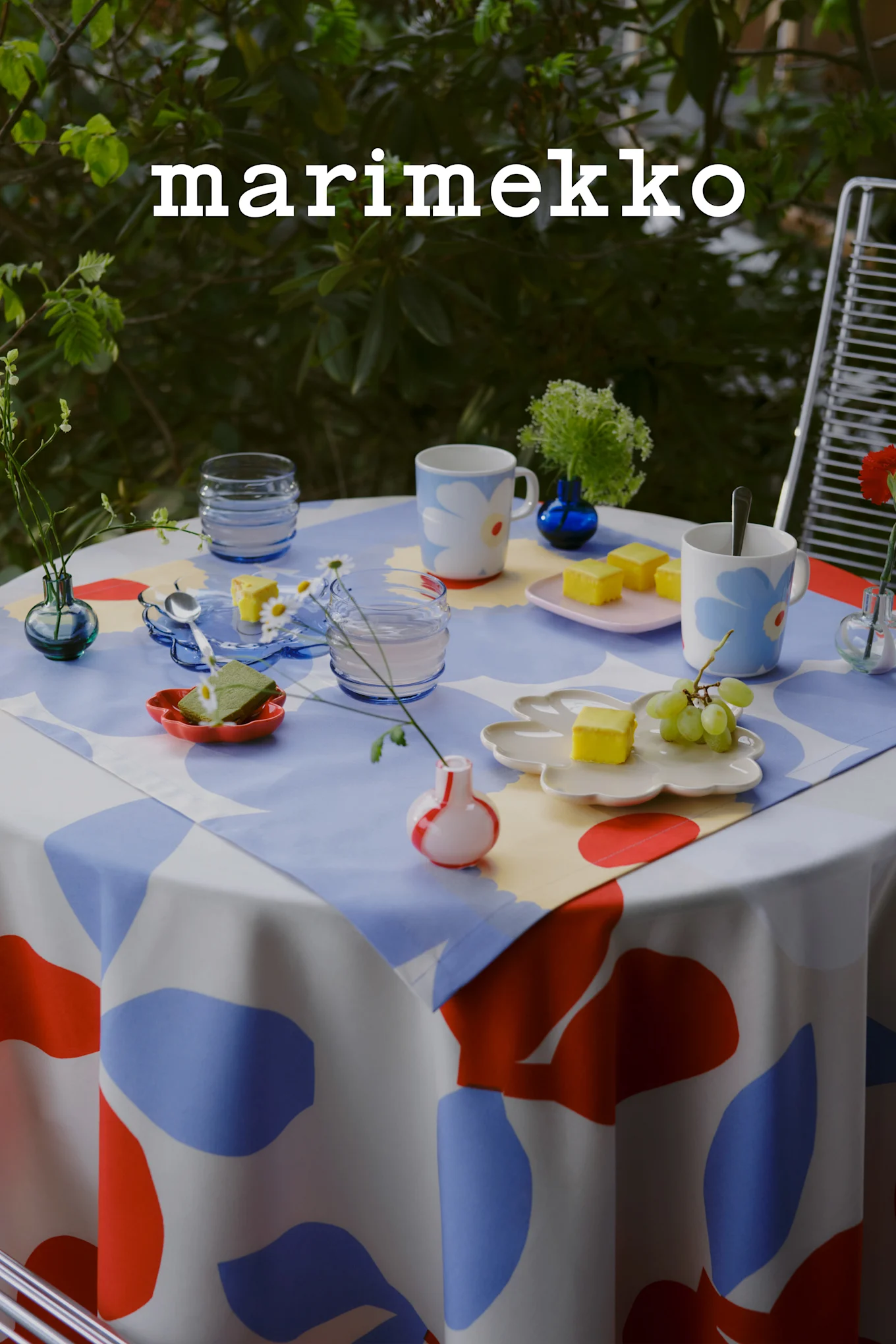 Marimekko logo. An outdoor table set with a blue, red, and white abstract pattern tablecloth, floral mugs, yellow cakes, grapes, and small vases.