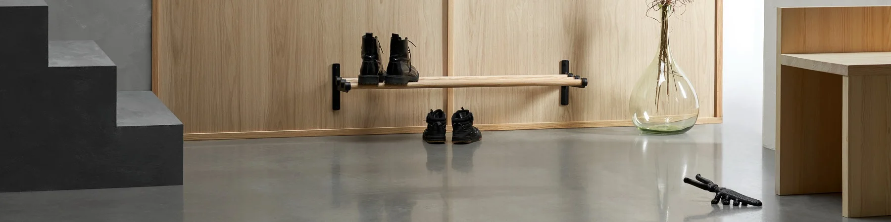 Entryway with a wooden shoe rack holding black boots, a large glass vase, and a decorative black animal figure on a concrete floor.