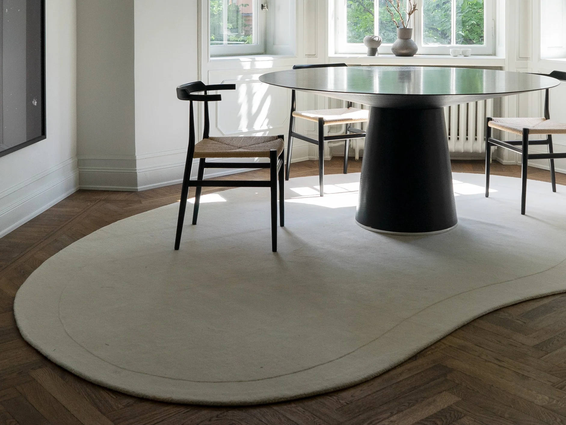 Modern dining room with a black round table, black chairs with woven seats, a cream irregular rug on a herringbone floor.