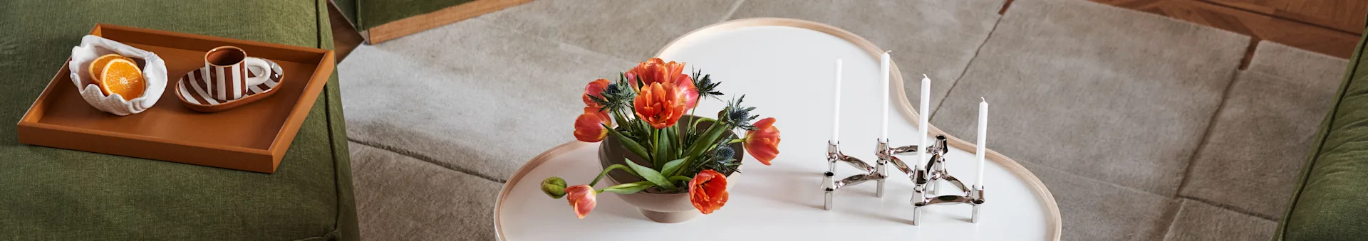 Green sofa, coffee table with orange tulips and silver candelabra. Tray on sofa has orange slices and striped cup.