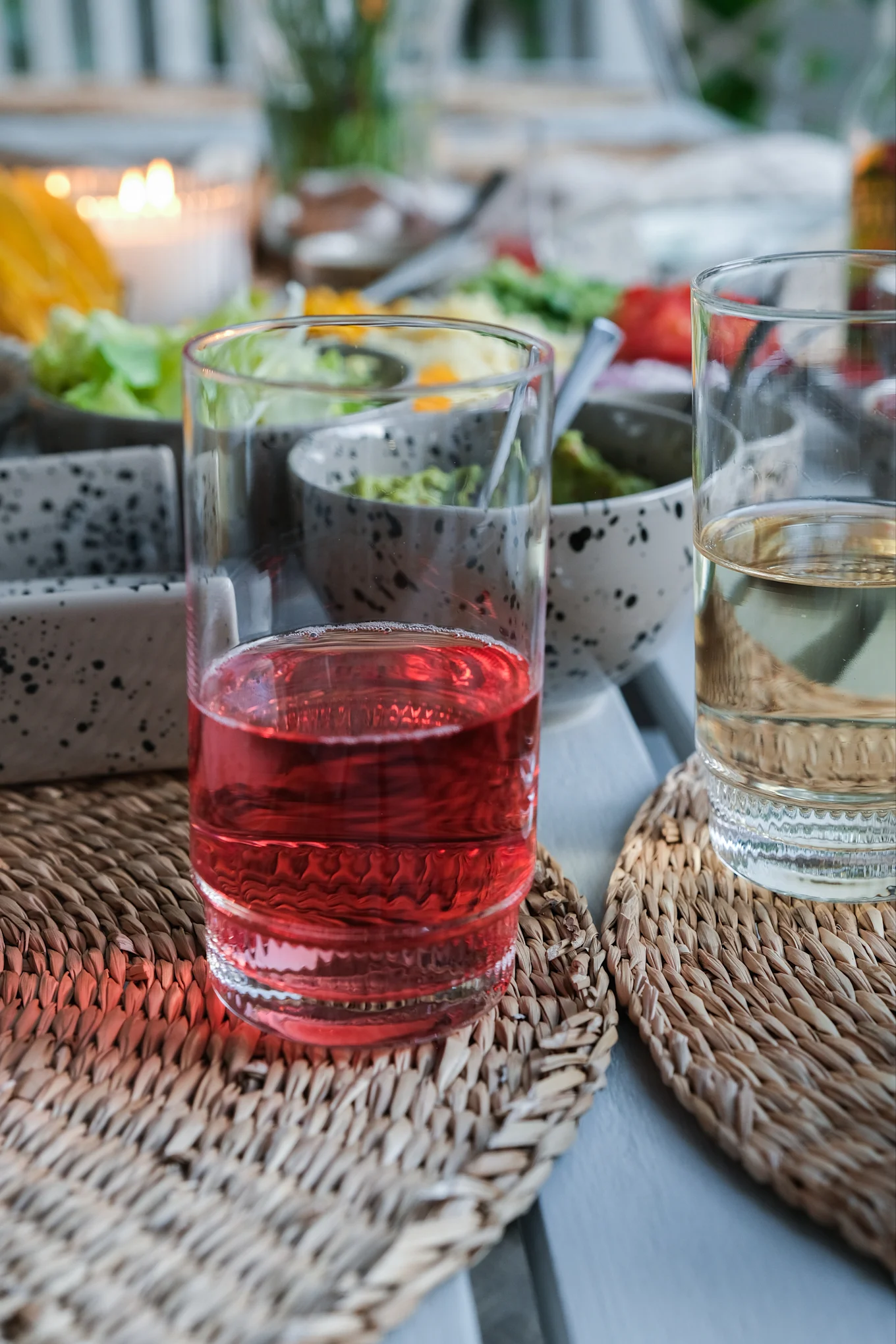 Two glasses, one with red drink and one with light drink, on woven placemats at a table with food and a candle.