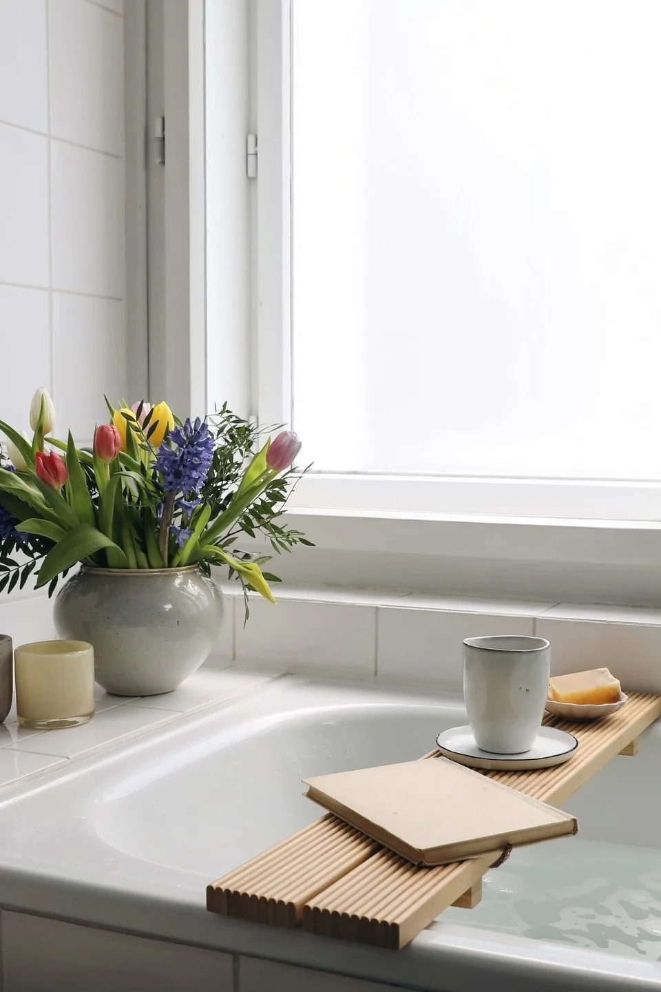 A cozy bathroom scene with a bathtub, a wooden bath caddy holding a book, mug, and soap, alongside a vase of colorful flowers.