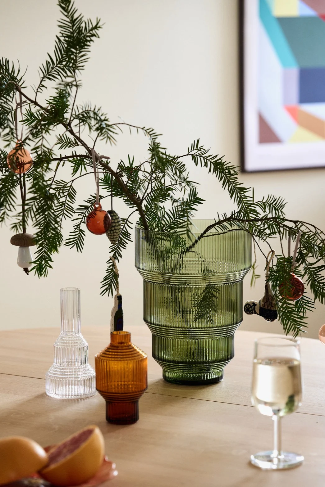 Decorated pine branches in a green ribbed vase, with clear and amber vases, a wine glass, and grapefruit on a wooden table.