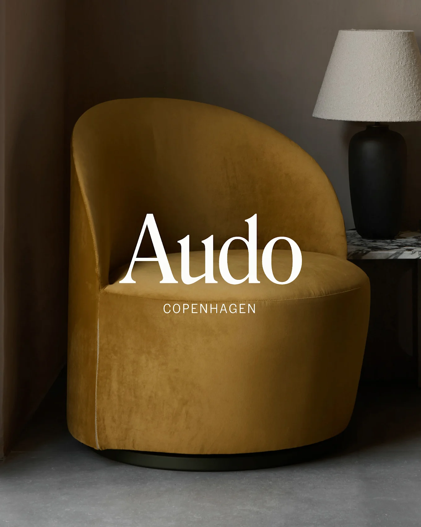 Mustard yellow velvet armchair with 'Audo Copenhagen' text overlay, beside a black lamp on a marble table.