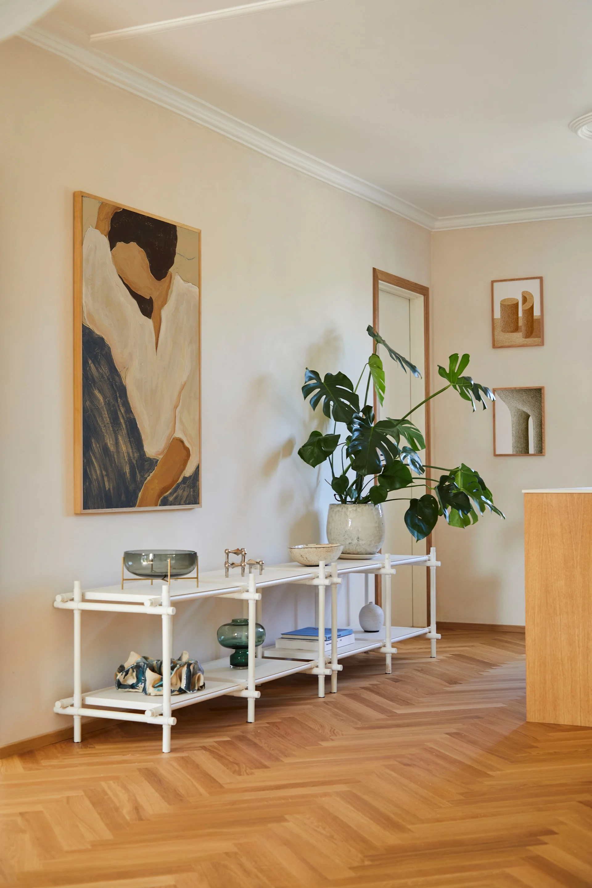 A bright room with a white shelving unit, a large Monstera plant, an abstract painting, and a herringbone wooden floor.