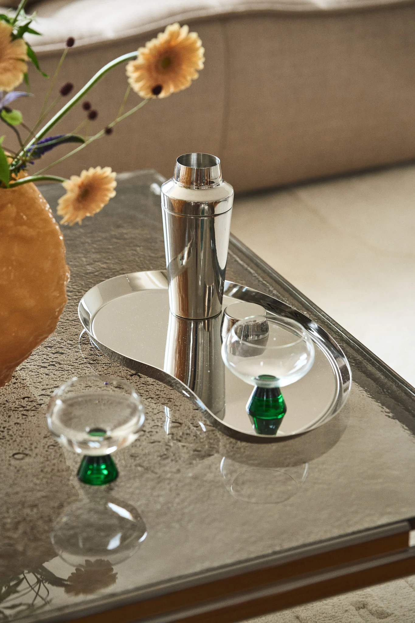 A silver cocktail shaker, an organic-shaped silver tray, and two clear glasses with green bases on a glass table.