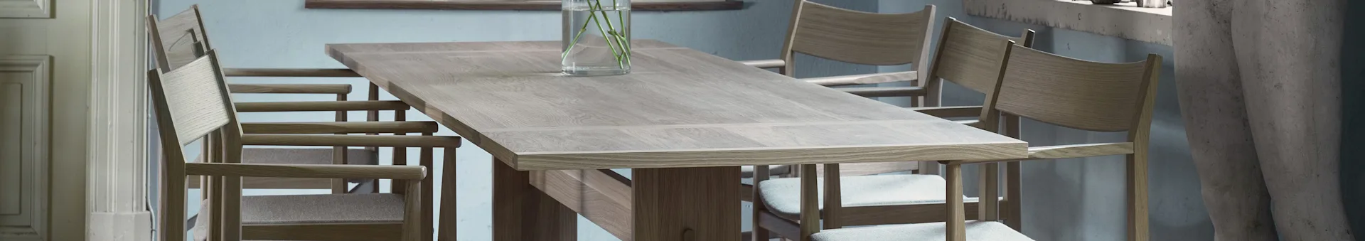 Light wooden dining table with matching chairs and a glass vase holding green stems.