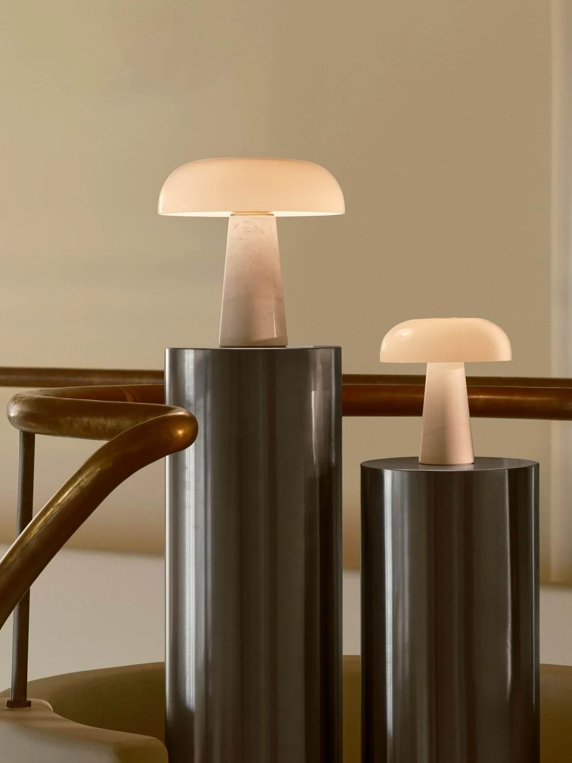 Two glowing mushroom-shaped table lamps, one large and one small, on cylindrical pedestals next to a brass railing.