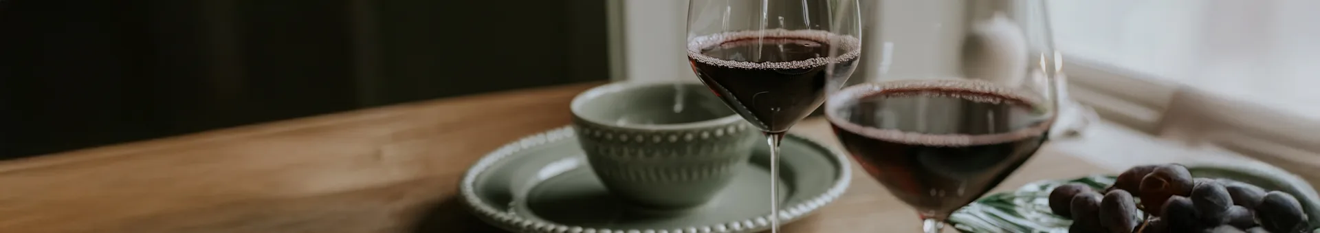 Two glasses of red wine on a wooden table, next to a green bowl and dark grapes.