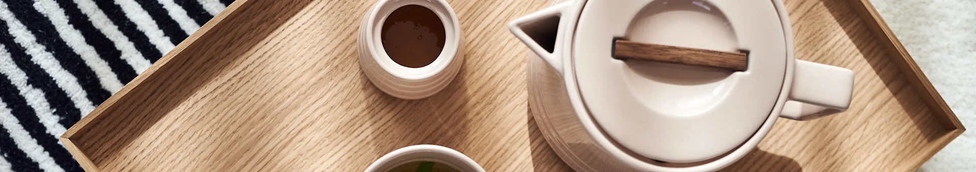 Overhead view of a light wooden tray with a beige teapot, a cup of dark liquid, and a cup of green liquid. A striped textile on the side.