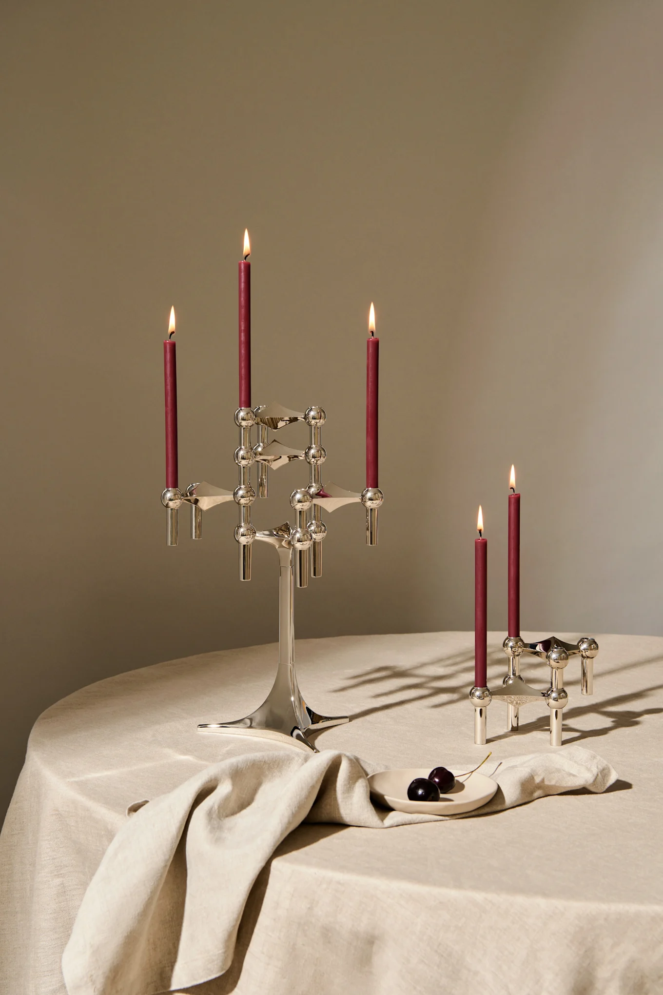 A large silver candelabra with four lit red candles and a smaller matching two-candle holder on a linen table with dark cherries.