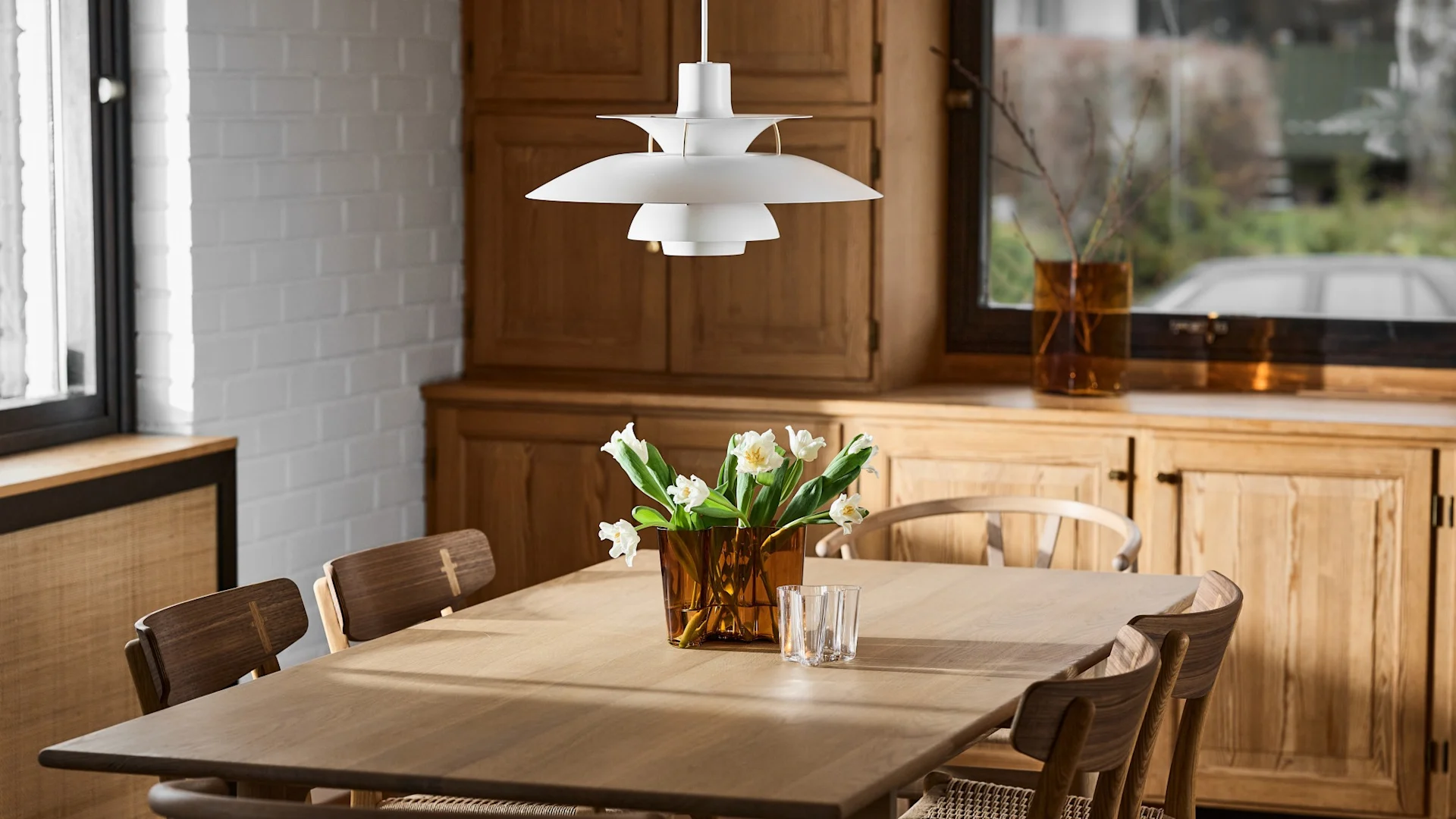 A white tiered pendant lamp hangs above a wooden dining table with an amber vase of white flowers and a clear glass.