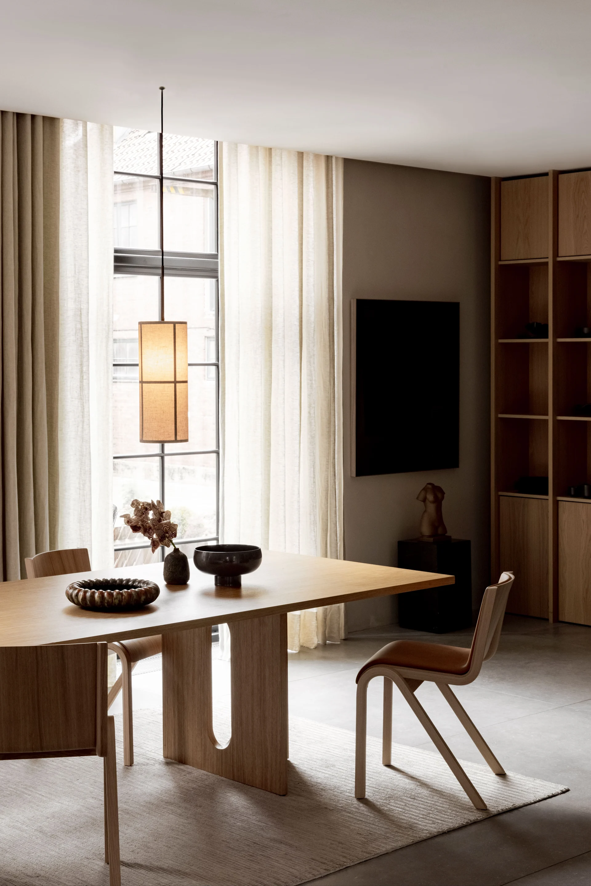 A modern dining room with a light wood table, two chairs, a dark decorative bowl, dried flowers, and sheer curtains.
