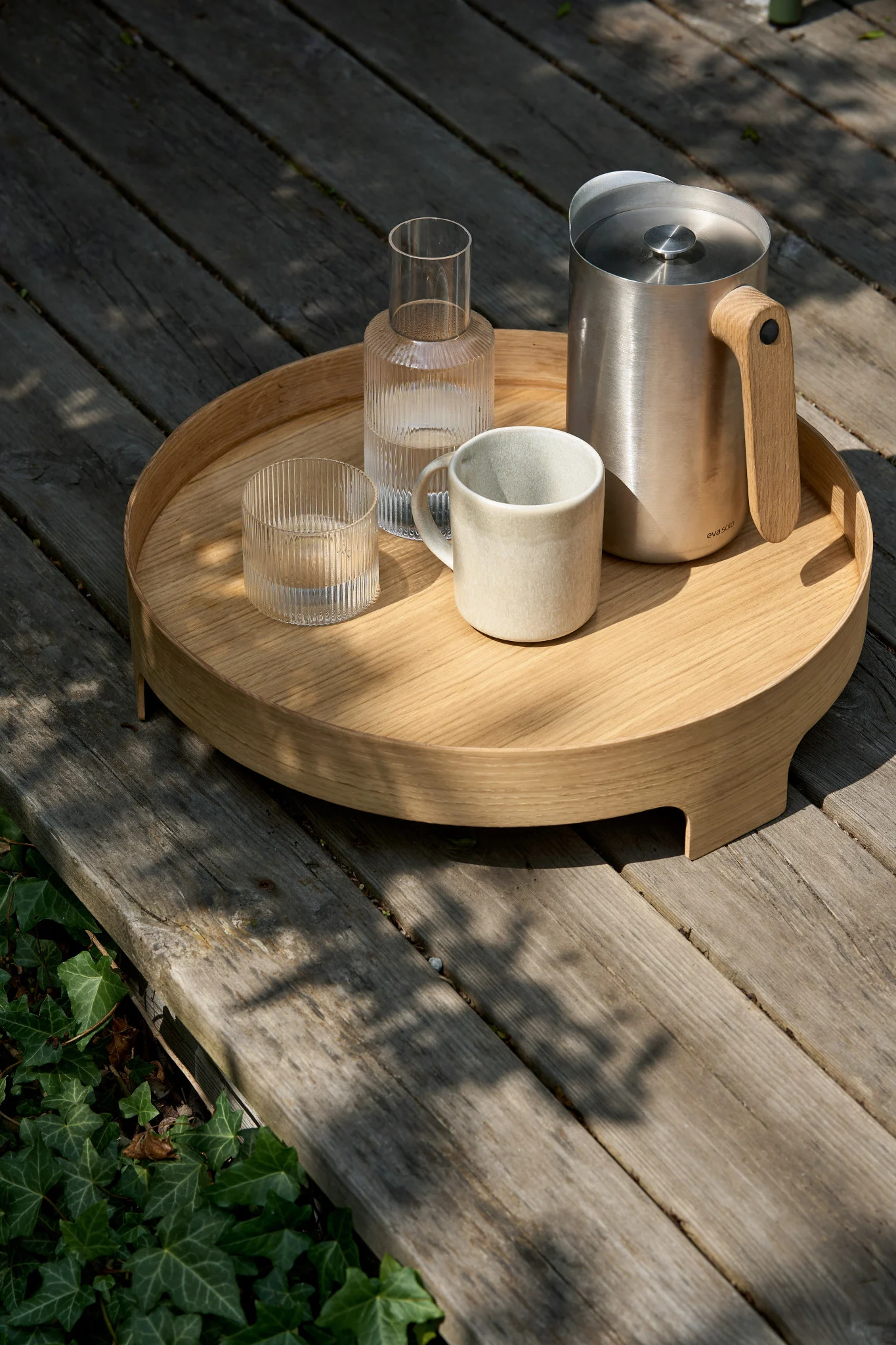 A round wooden tray on a deck holds a stainless steel pitcher with a wooden handle, a ribbed glass carafe, a ribbed glass, and a ceramic mug.