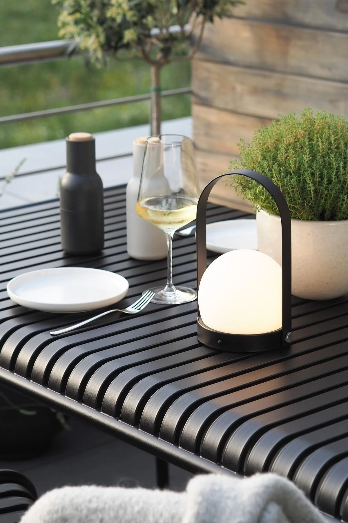 Glowing portable lantern on a black outdoor table, with white wine, plate, and green plant.
