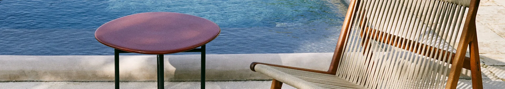 A round, reddish-brown side table and a lounge chair with a wooden frame and woven rope seating by a blue pool.