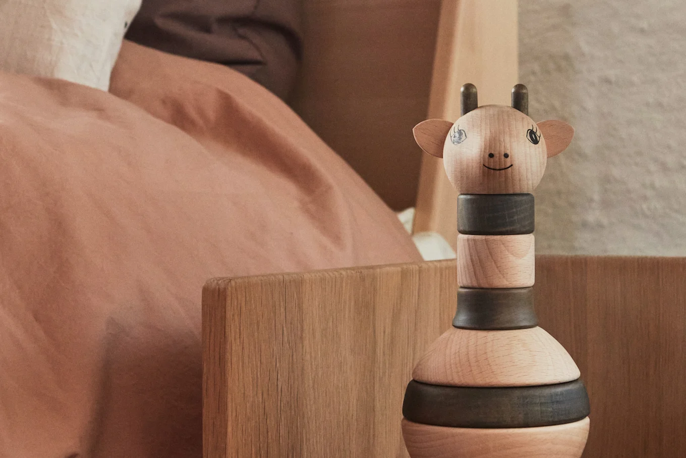 A wooden giraffe stacking toy with a smiling face stands next to a bed with pink bedding.