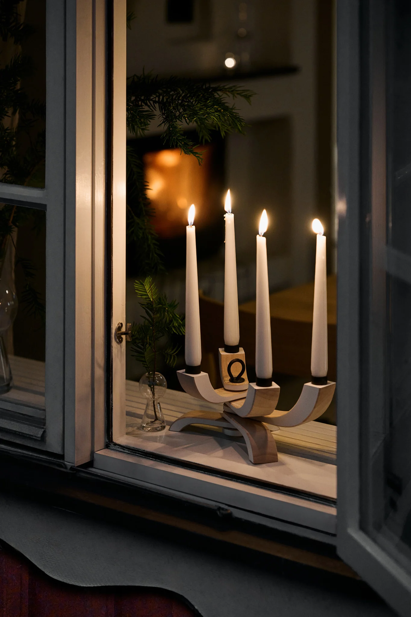 Four lit white candles in a wooden candle holder on a windowsill with evergreen branches, a warm fire visible indoors.