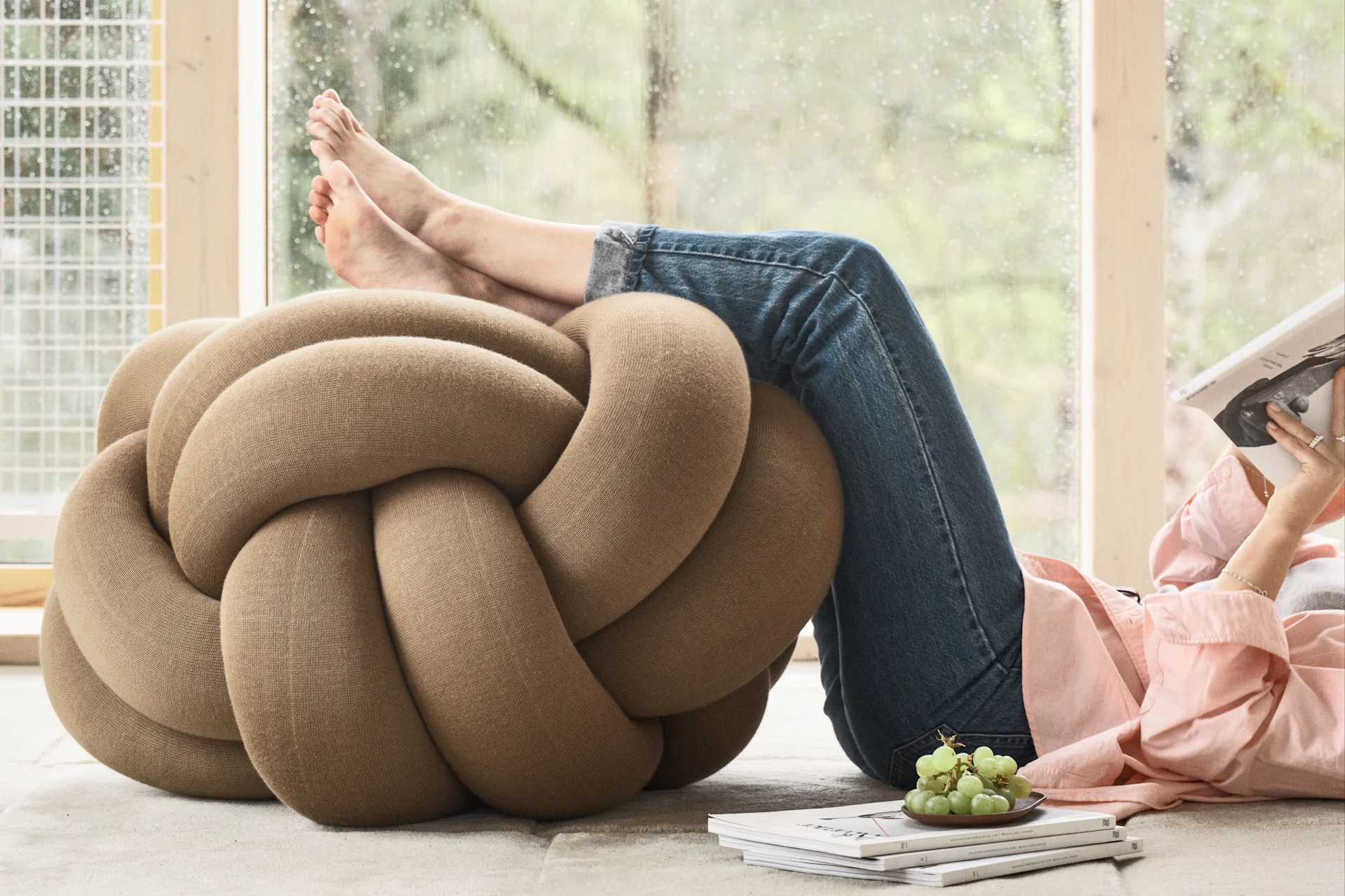 Person relaxing with feet on a large beige knot pouf, reading a book. Green grapes and magazines are nearby.