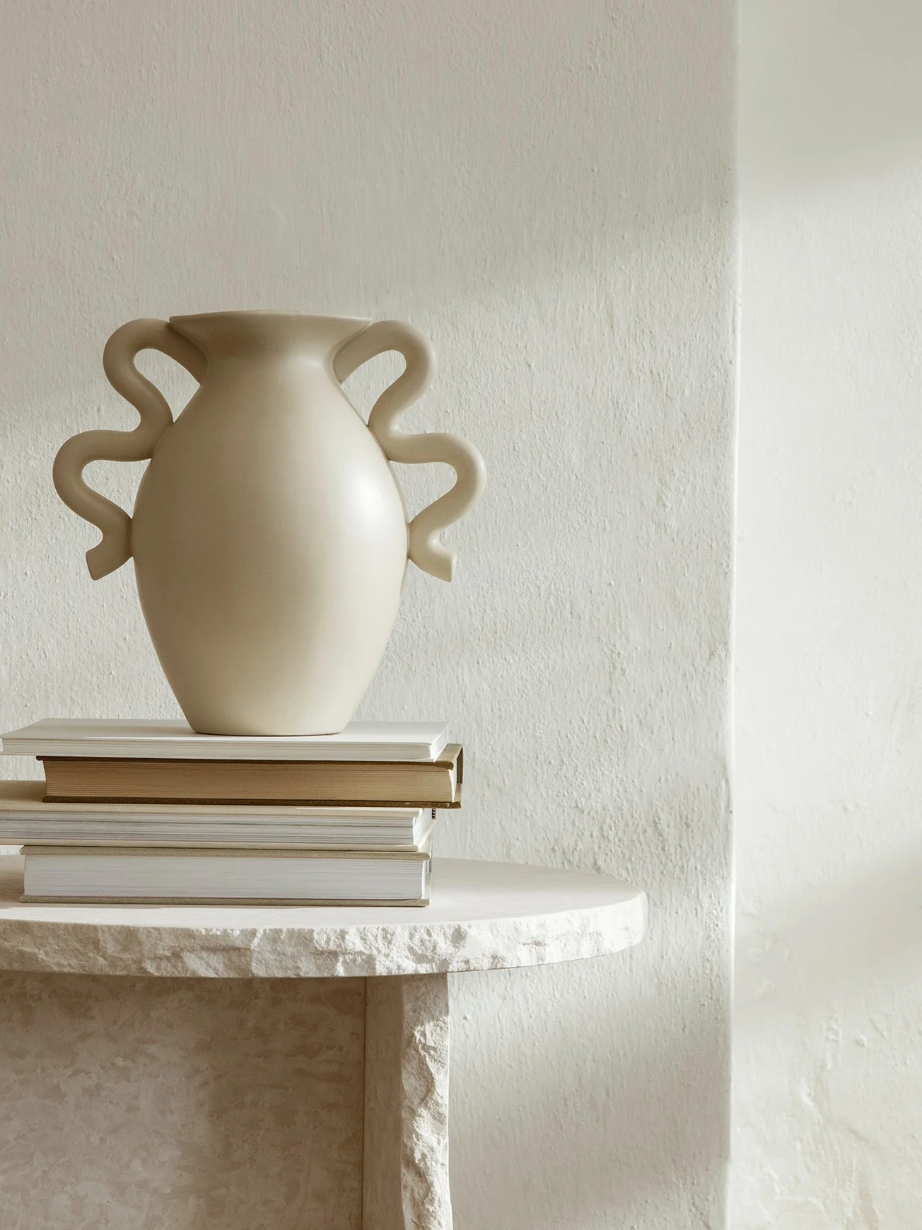 Off-white vase with wavy handles on a stack of books, resting on a light stone table in a minimalist setting.