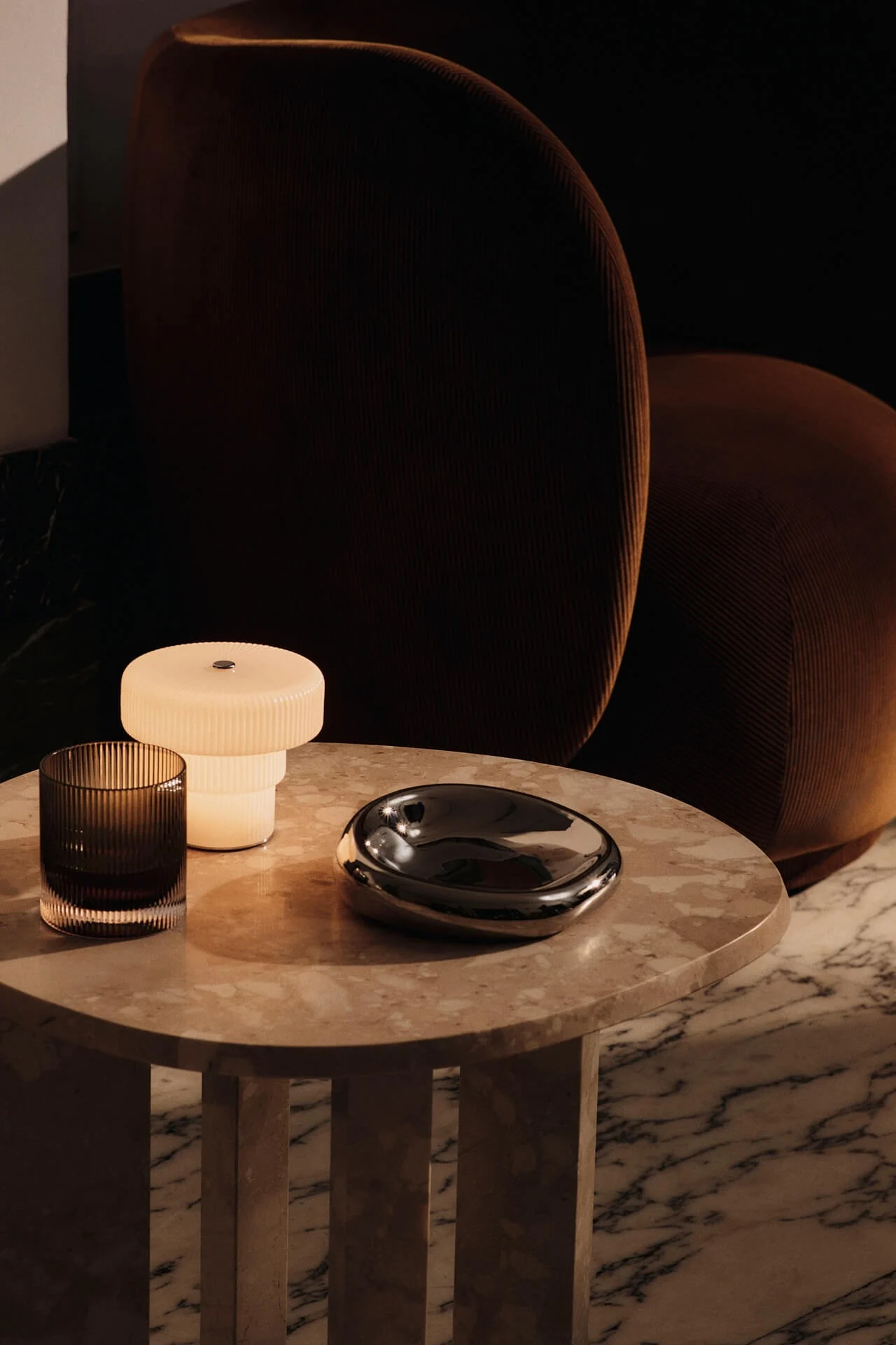 A glowing white ribbed lamp, a dark ribbed glass, and a silver organic dish on a light marble table, with a brown ribbed chair in the background.