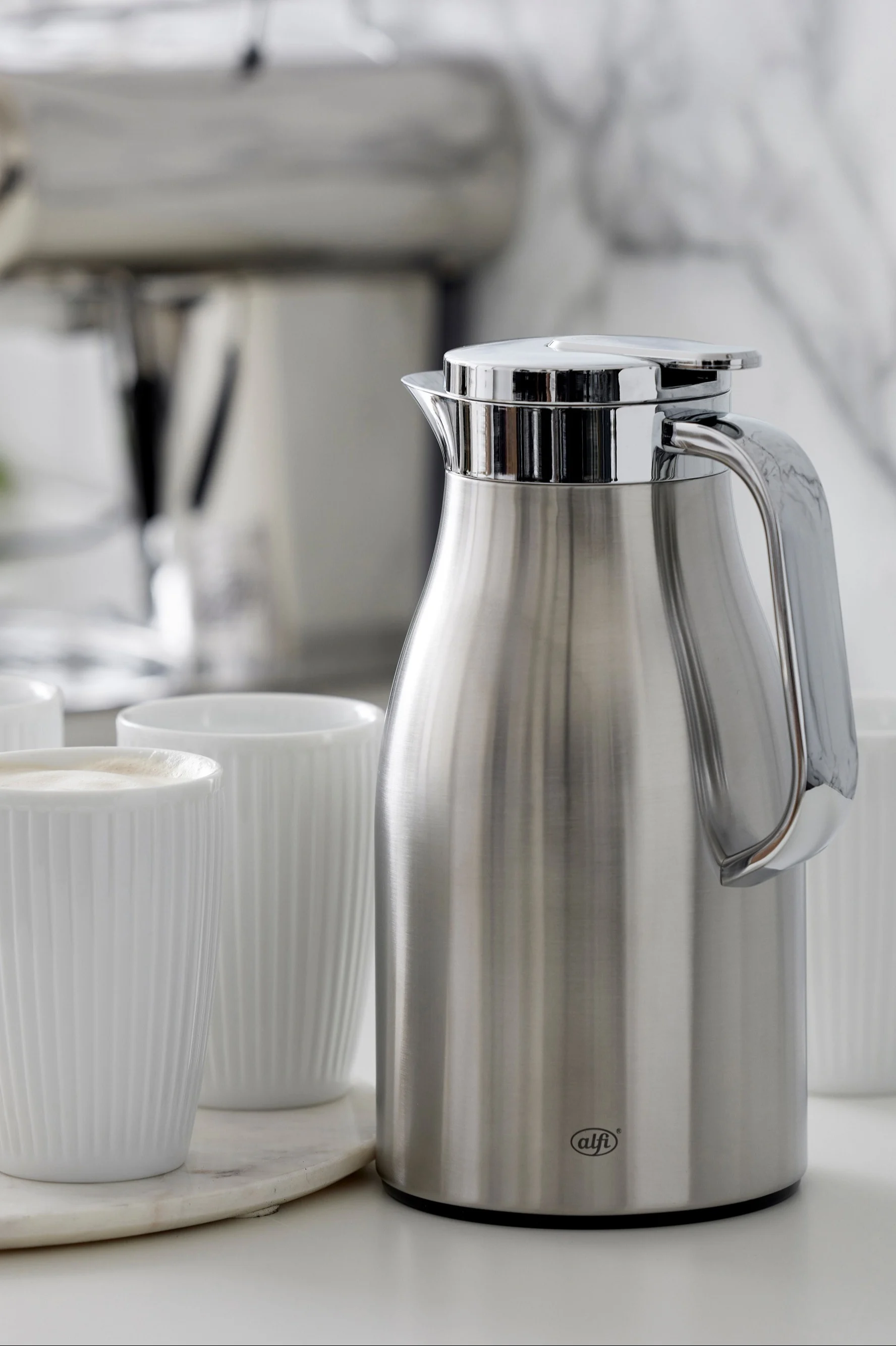 Alfi brushed steel thermal carafe with chrome lid, handle, and white ribbed mugs on marble tray.