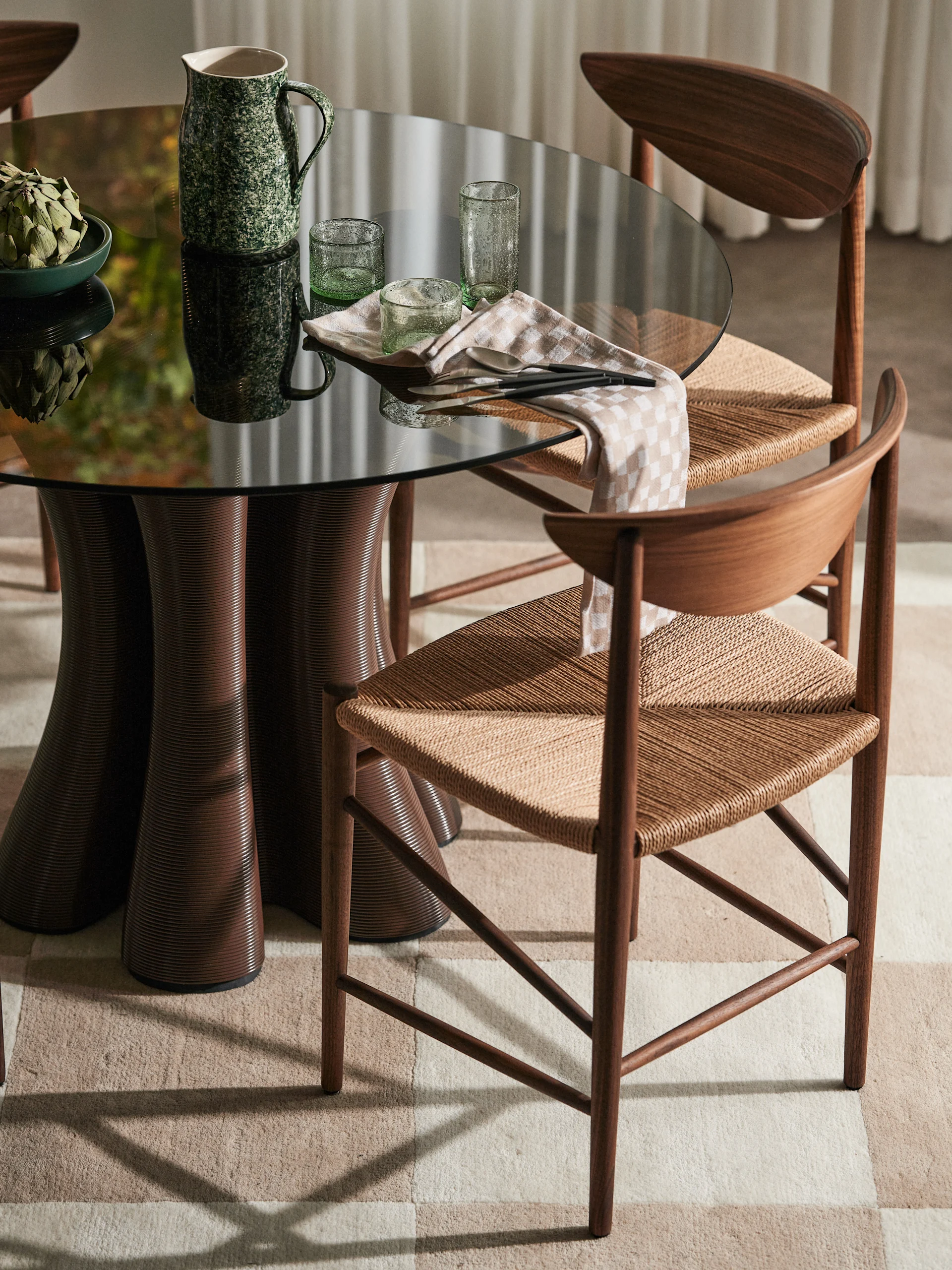 Dining table with dark glass top, fluted brown base, two wooden chairs with woven seats, green pitcher, artichoke, and glasses.