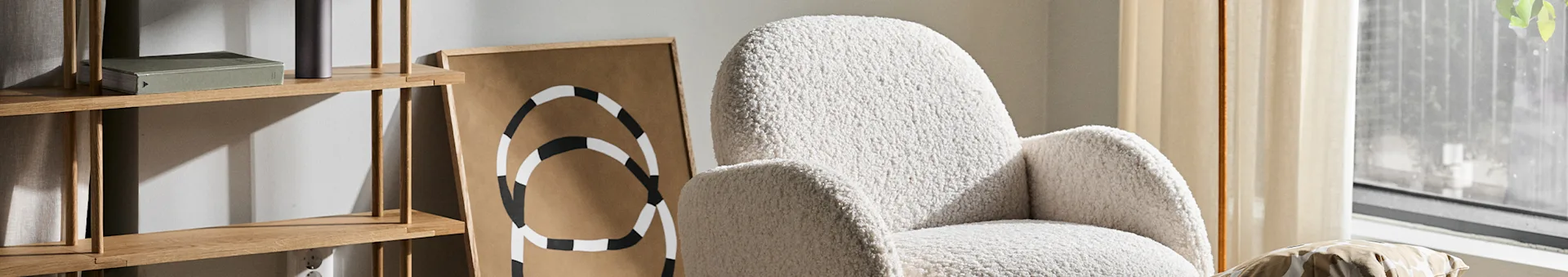 A fluffy white armchair, a wooden shelf with a book, and abstract art in a light-filled room.