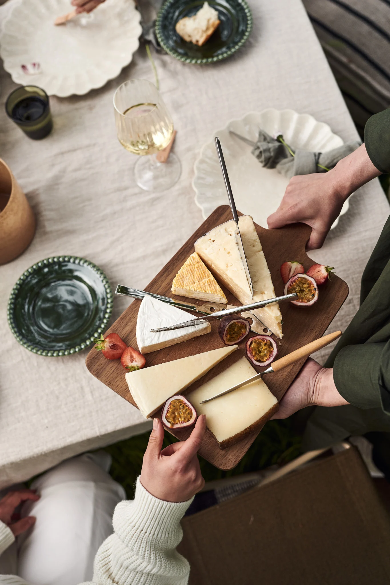 Hands serve a wooden cheese board with various cheeses, halved strawberries, and passion fruit on a table setting.
