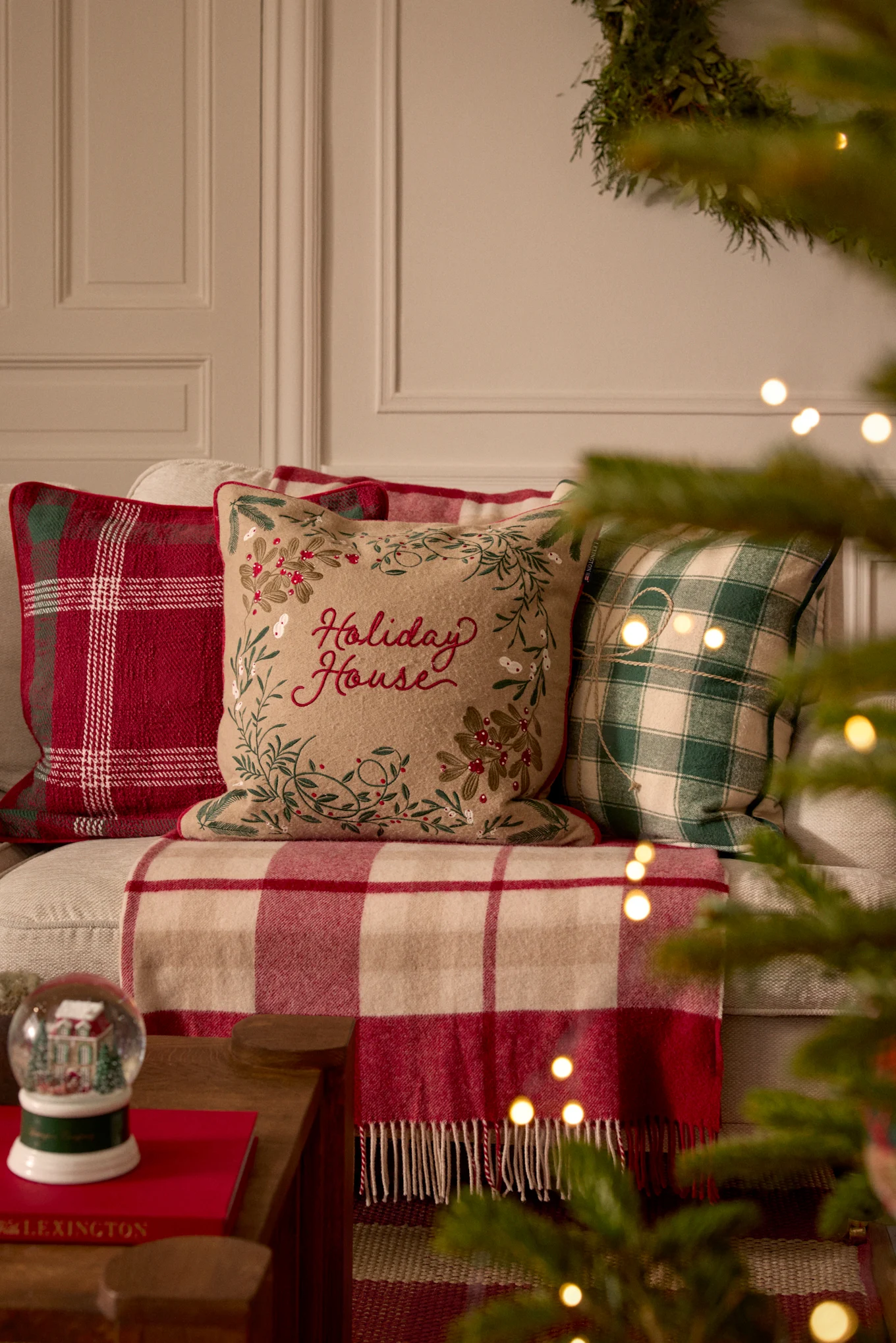 Three holiday pillows and a plaid blanket on a sofa, with a snow globe and Christmas tree lights nearby.