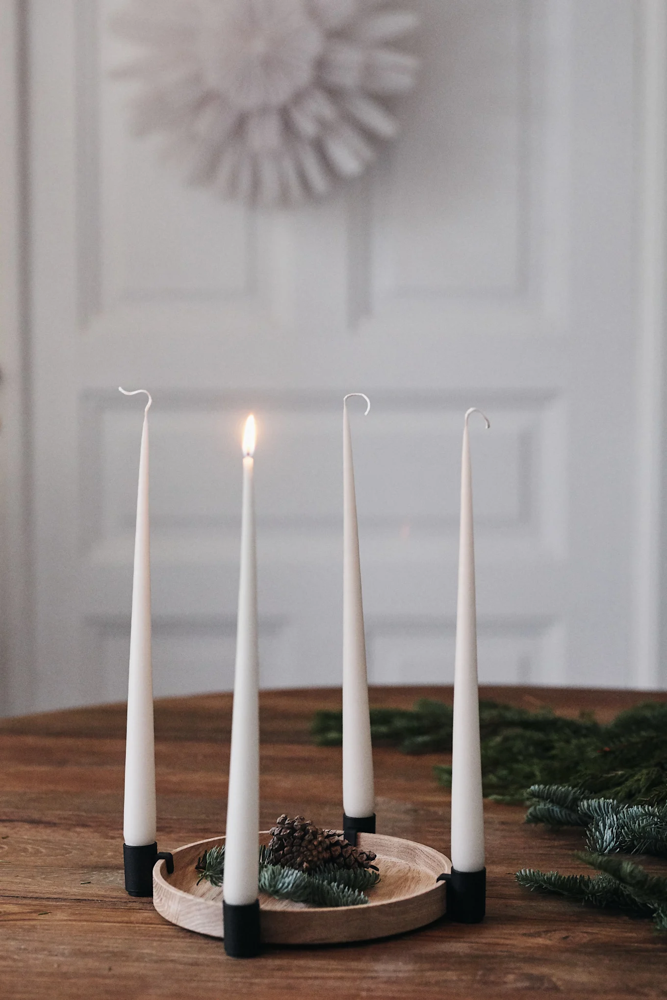 An Advent wreath with four cream tapered candles, one lit, arranged on a wooden tray with pinecones and fir branches.