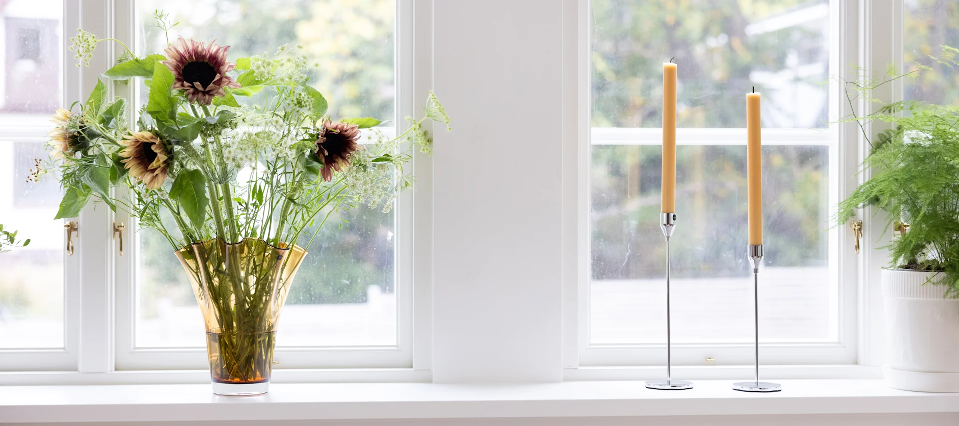 A bright window sill with a vase of sunflowers, two yellow candles in silver holders, and a potted green fern.