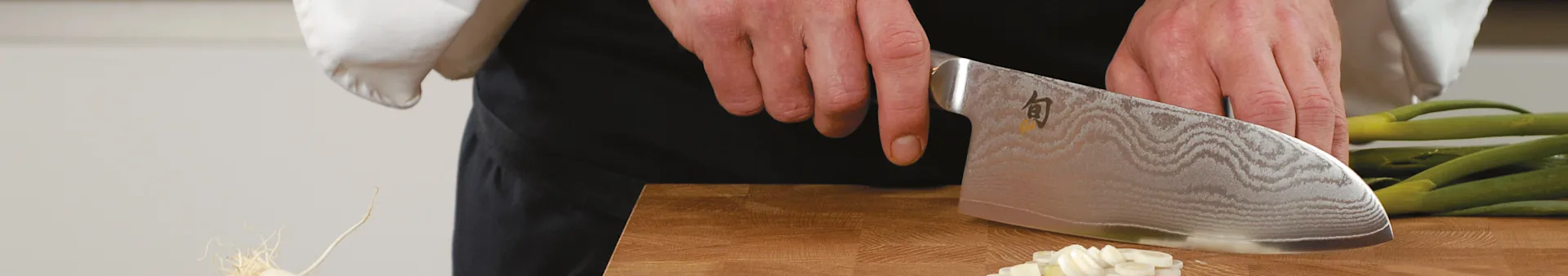 A chef's hands chop green onions on a wooden board with a large, patterned knife.