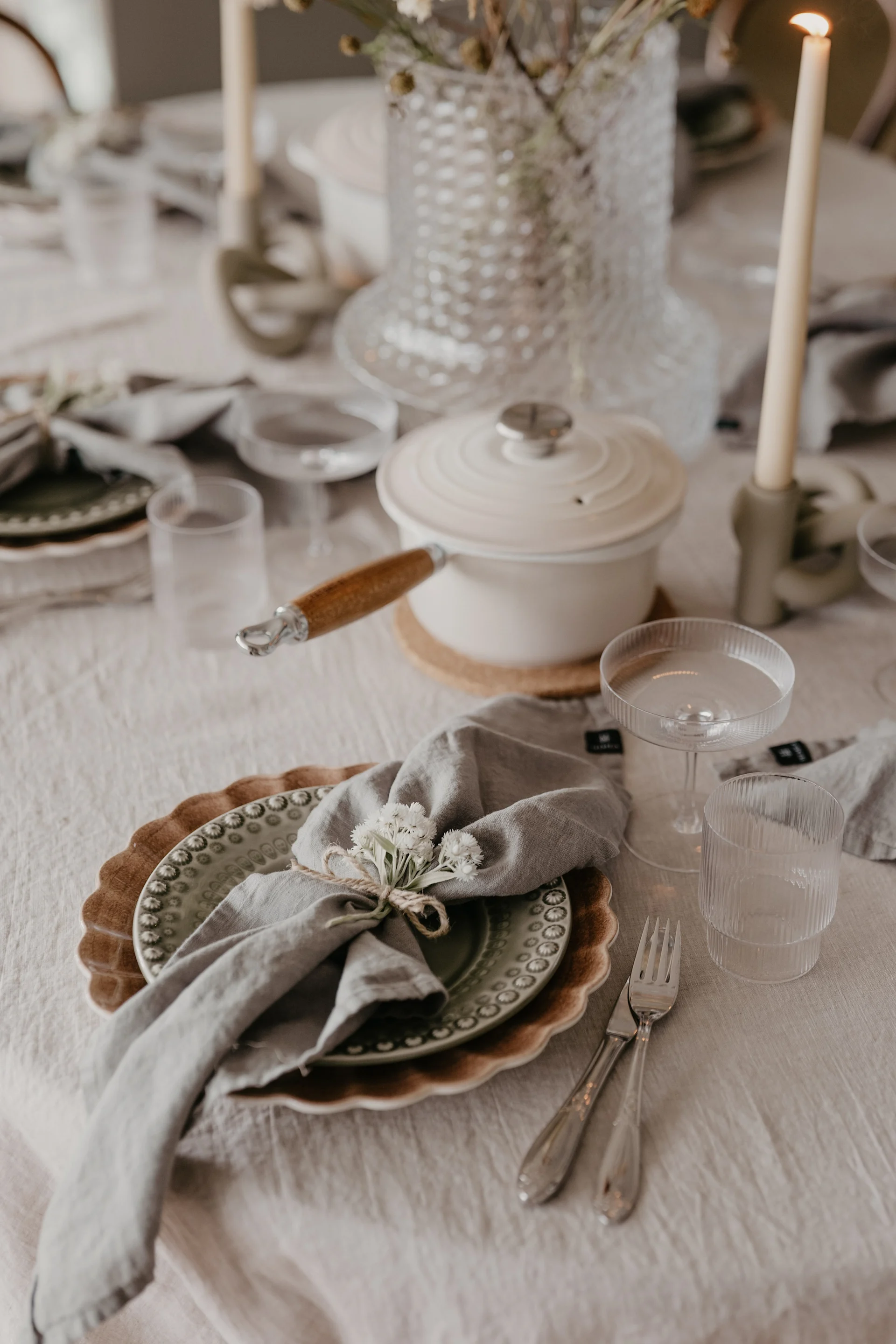 A festive dining table with a green plate, gray napkin tied with twine and white flowers, silverware, and a white pot.