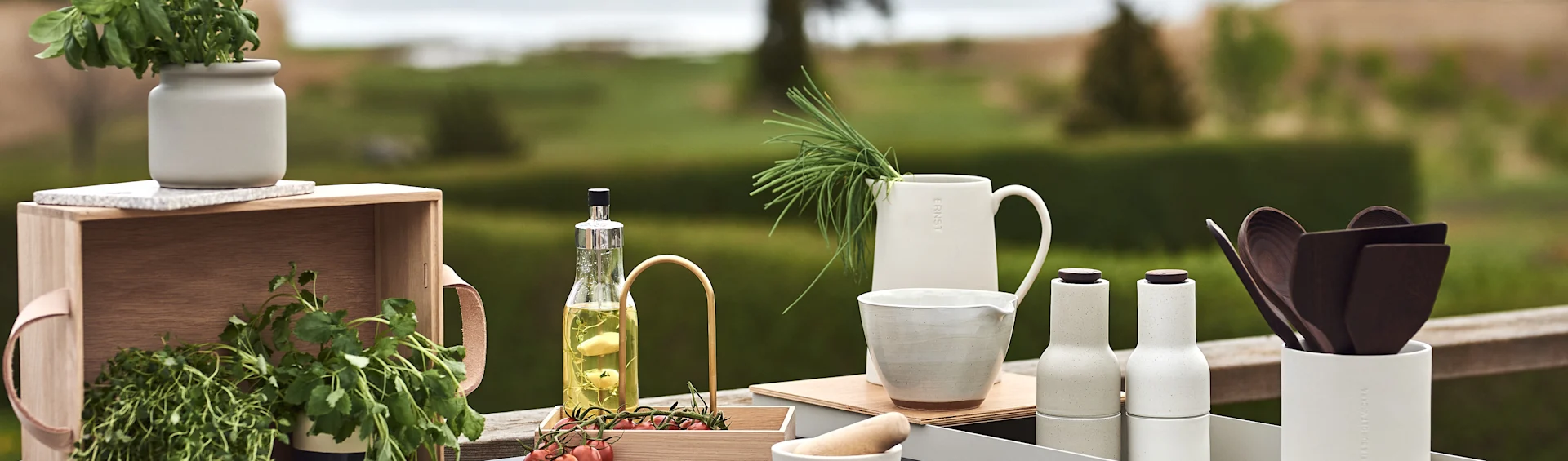 Assortment of ceramic kitchenware, fresh herbs, olive oil, and cherry tomatoes on a wooden surface outdoors.