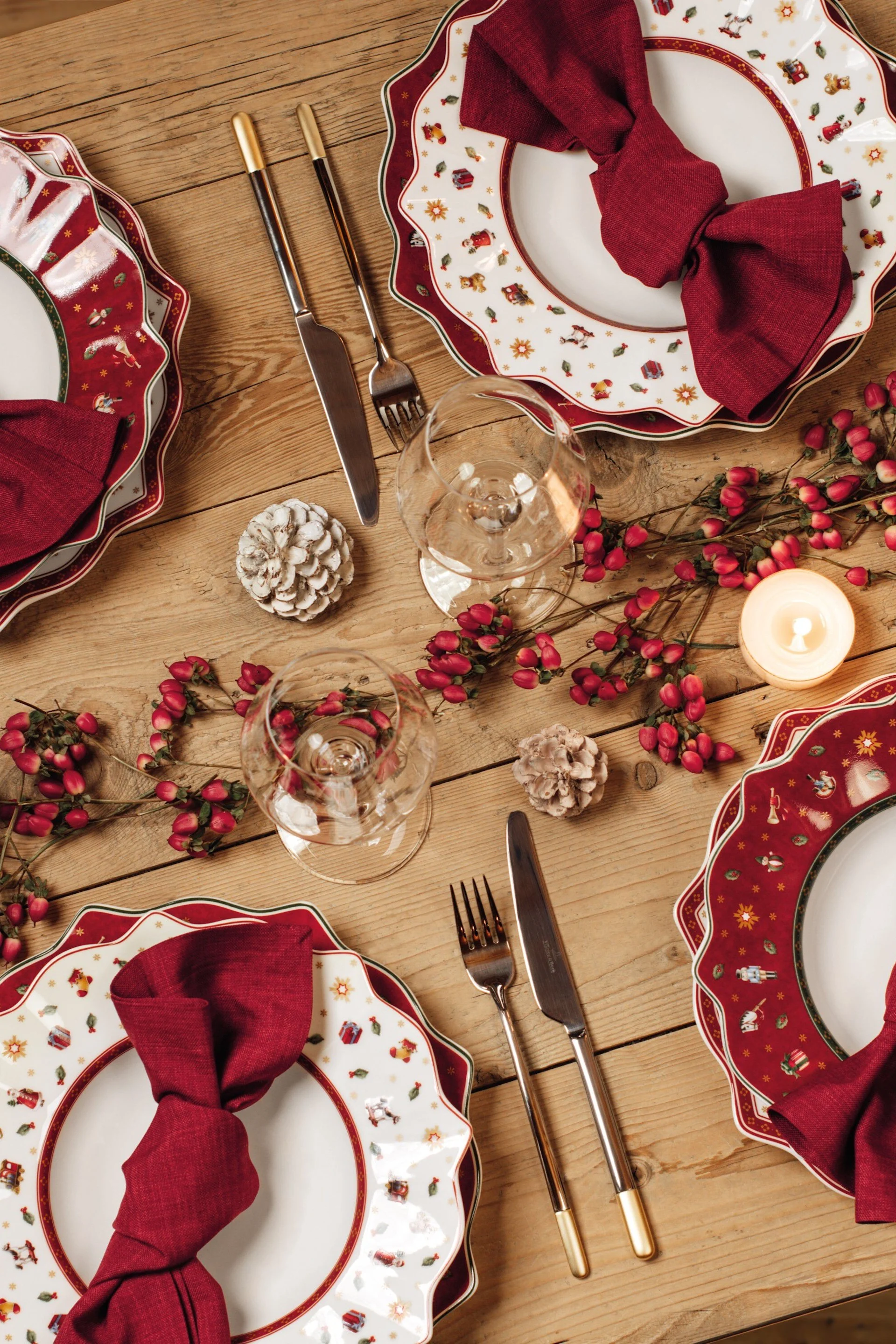 Festive wooden table set with four plates featuring Christmas toy patterns, red napkins, silverware, glasses, berries, and candles.