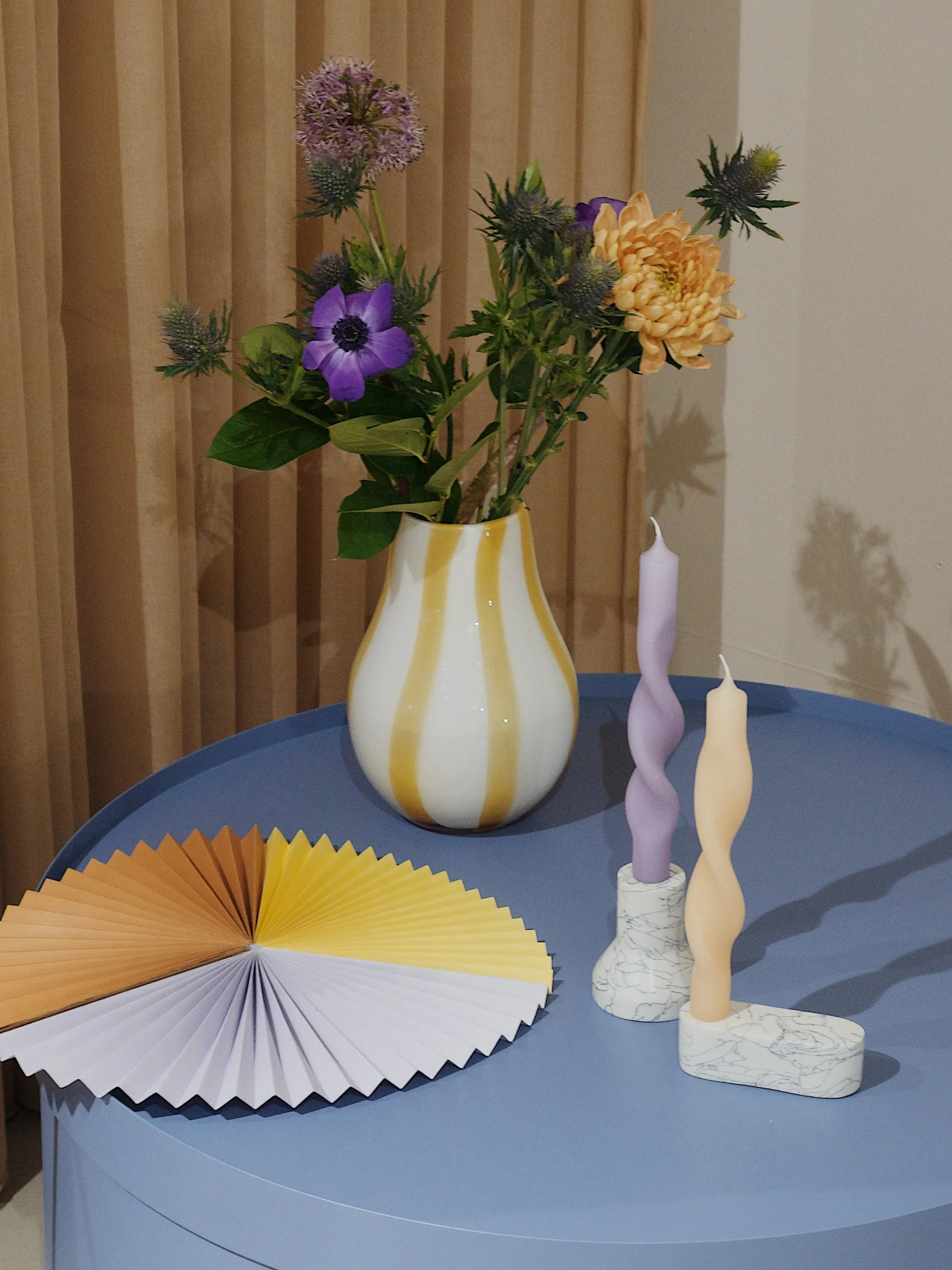 Floral arrangement in a striped vase, twisted candles in marble holders, and a colorful fan on a blue table.