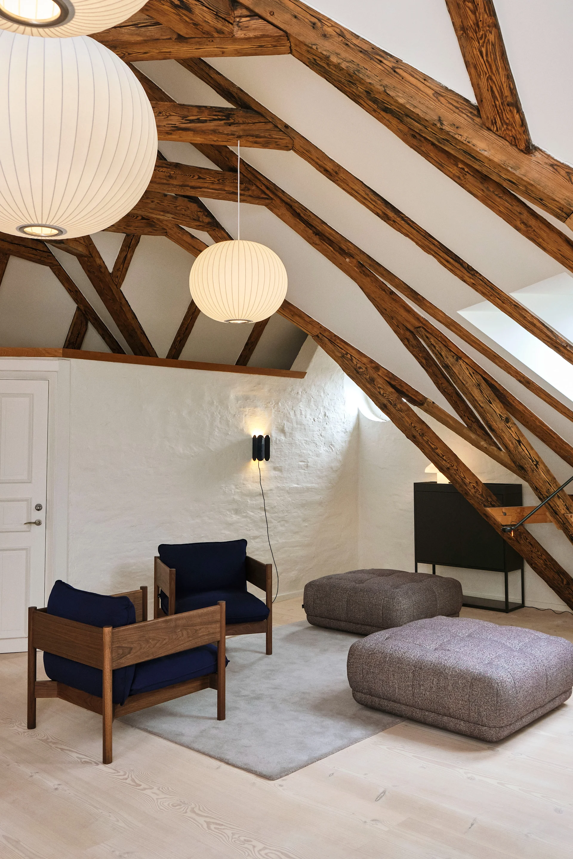 An attic room with exposed wooden beams, two round pendant lamps, blue chairs, grey ottomans, and a wall sconce.