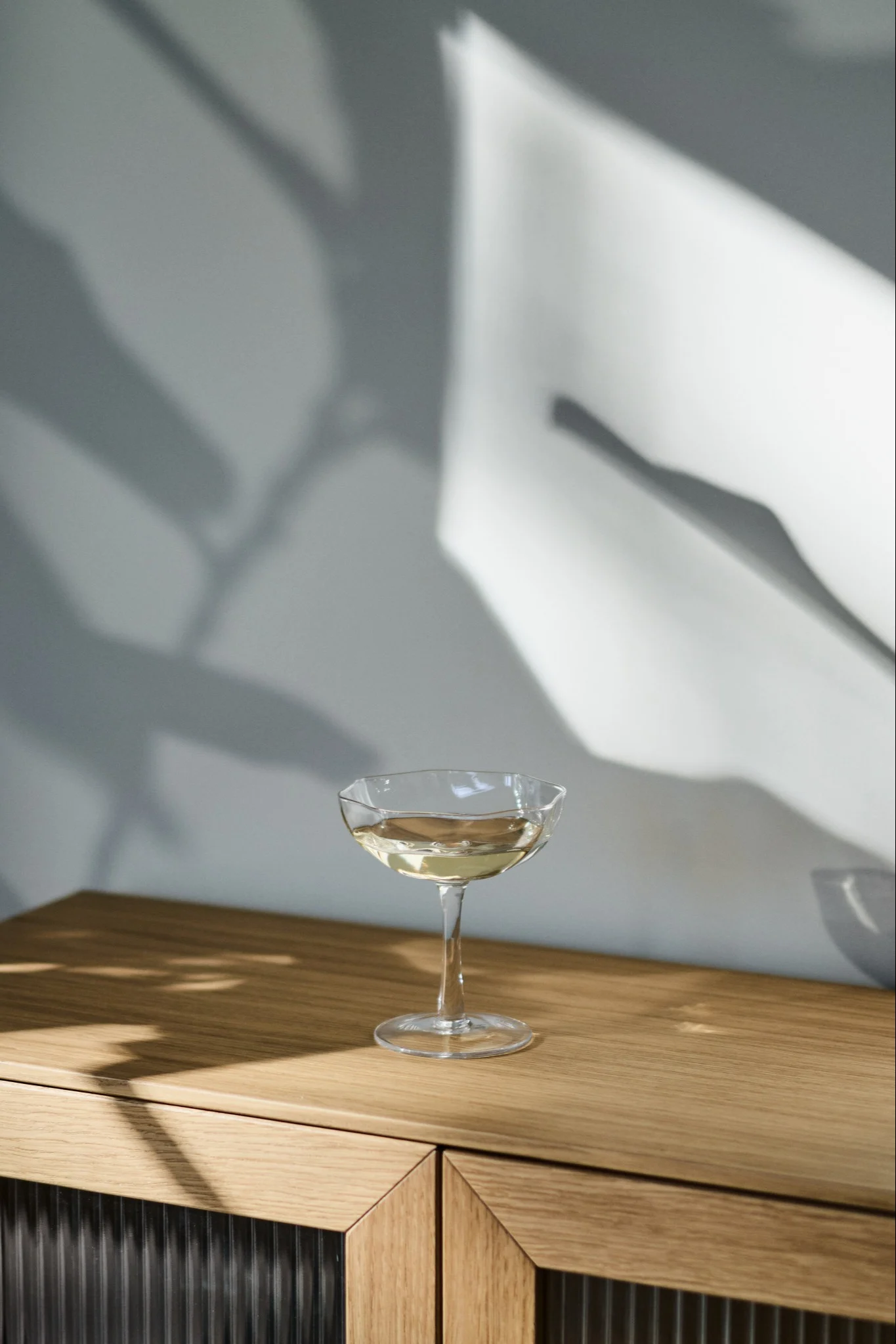 A coupe glass with a light drink on a wooden cabinet, with abstract shadows on the wall.
