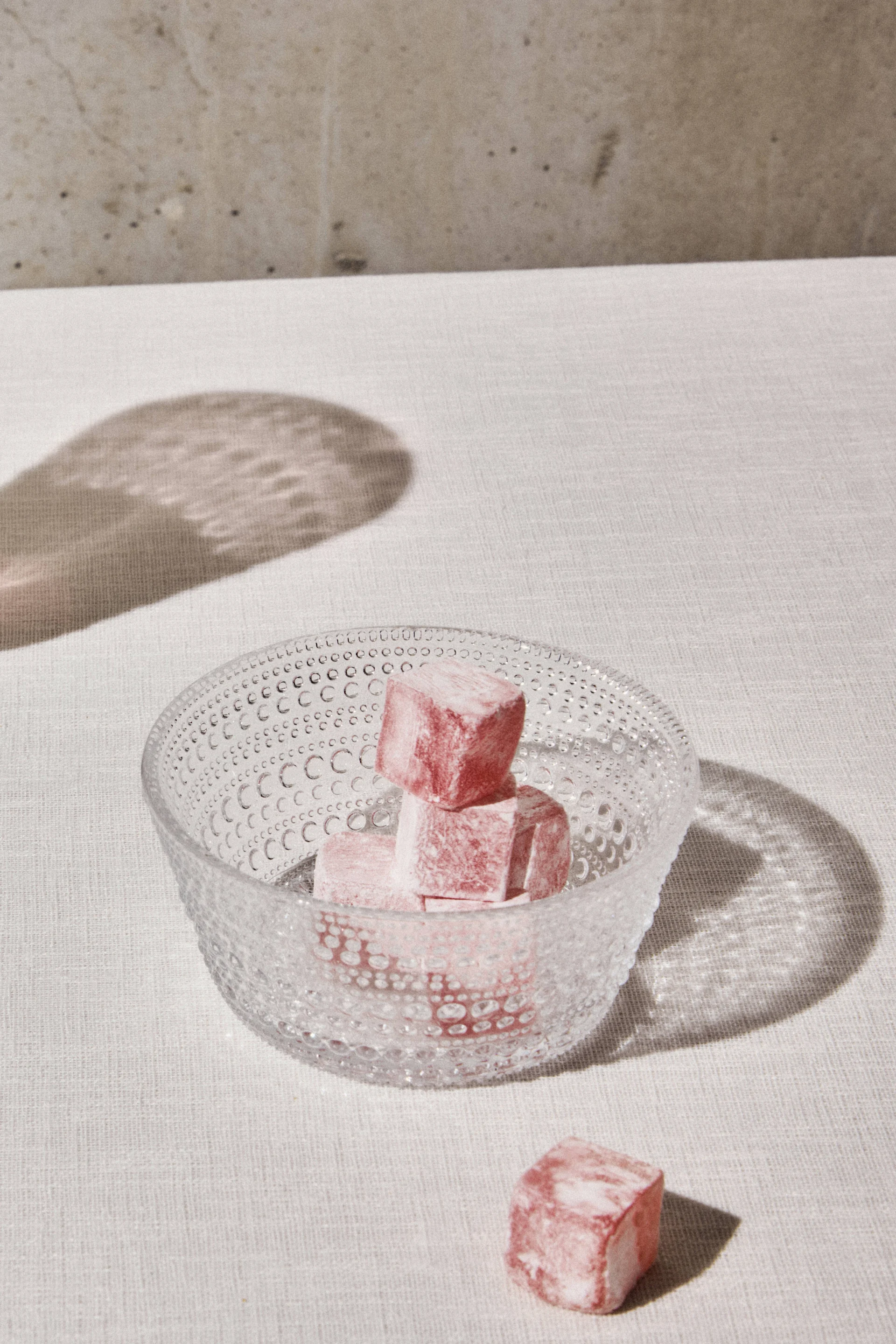 Pink Turkish delights in a clear textured glass bowl, with one delight outside on a light fabric surface.