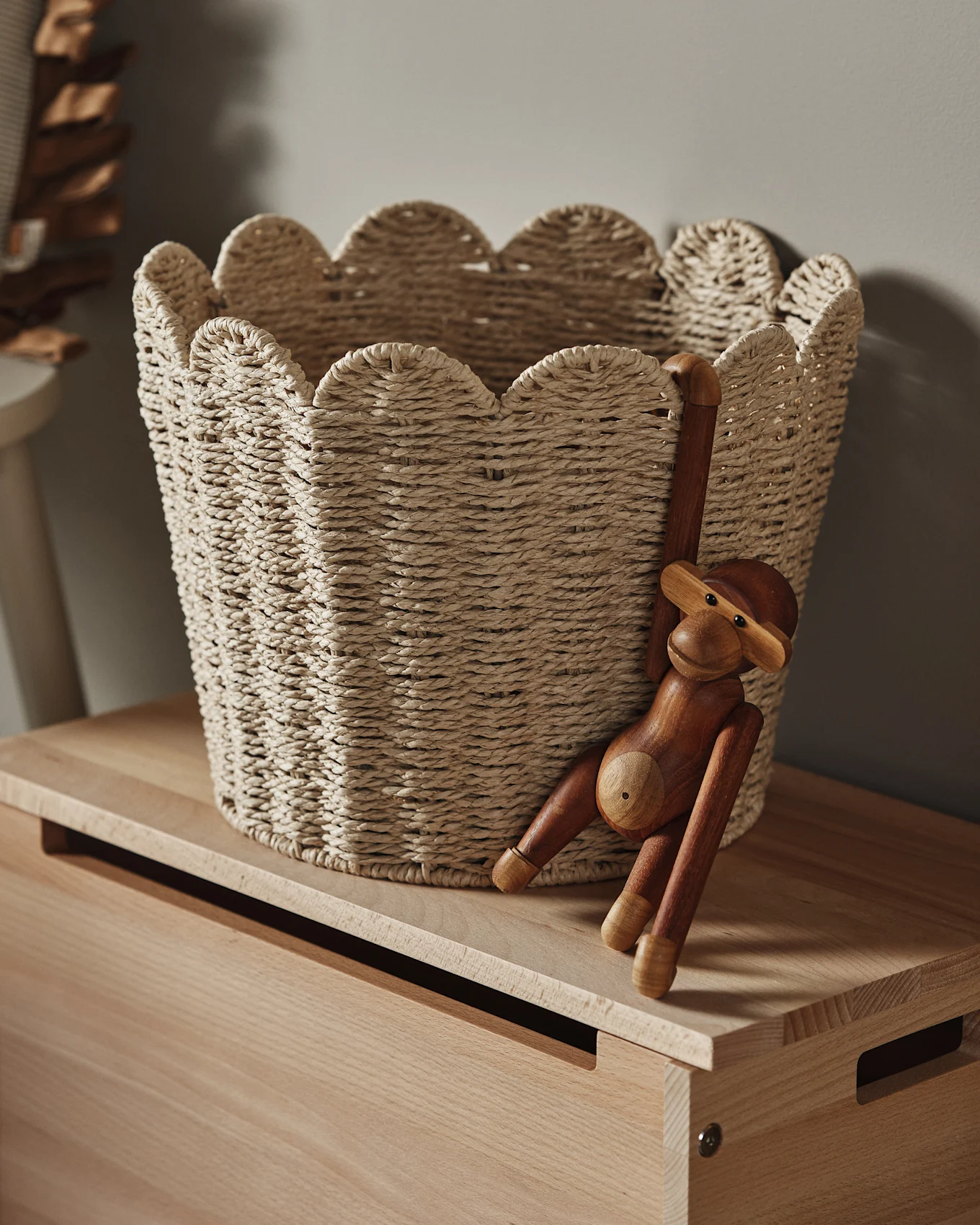 A light woven basket with a scalloped edge sits on a wooden surface, with a wooden monkey figurine hanging from its side.