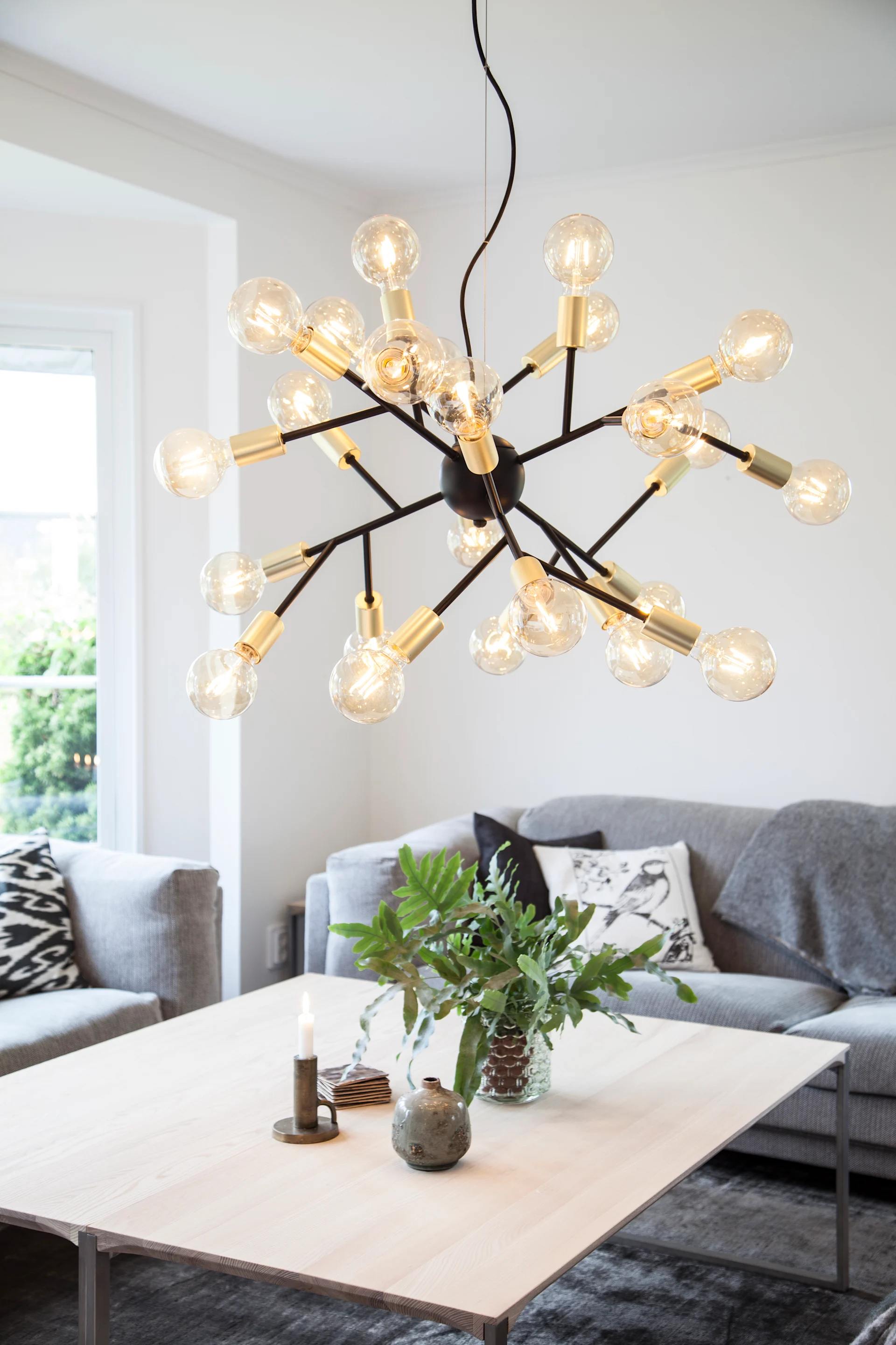 A modern sputnik-style chandelier with many glowing bare bulbs illuminates a living room with a light wood coffee table and grey sofa.