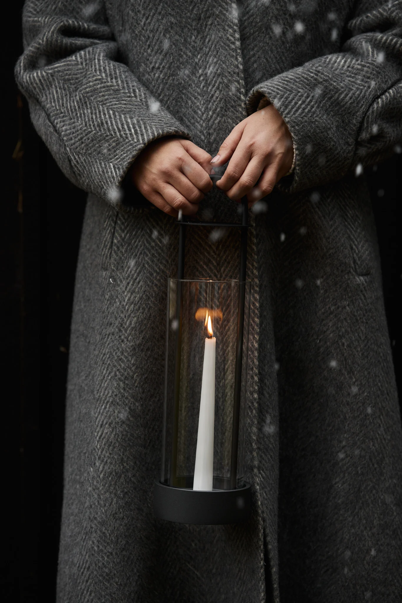 Person in a grey herringbone coat holding a glass lantern with a lit white candle in the snow.