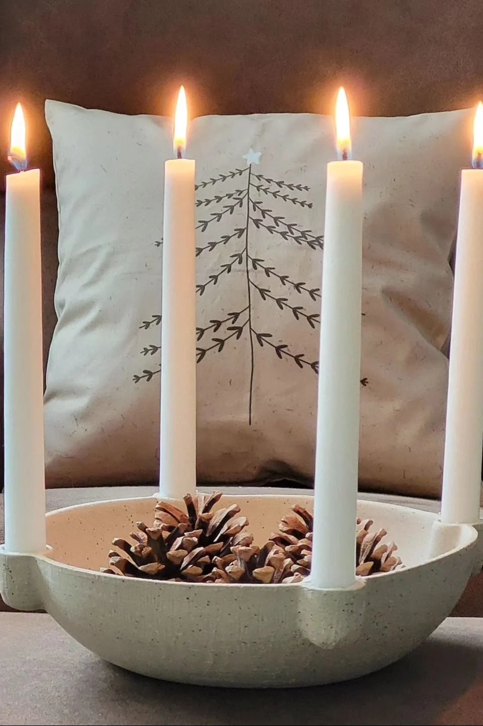 Four lit white taper candles in an off-white bowl with pinecones, against a pillow with a black outline Christmas tree design.